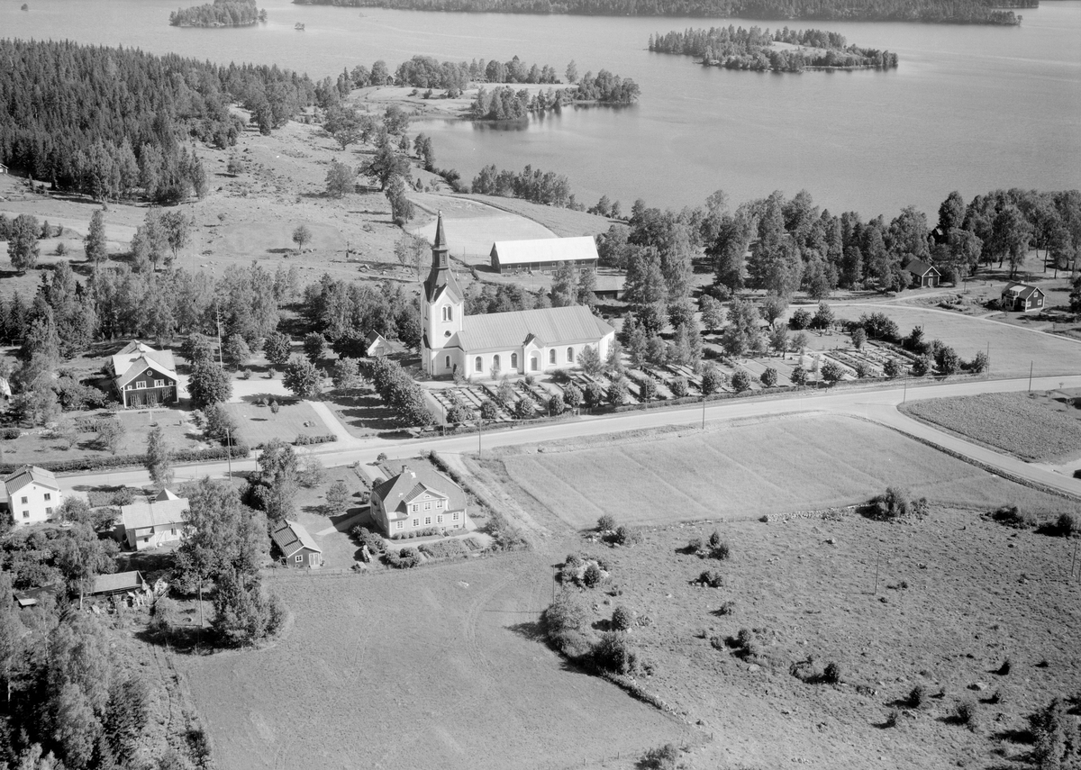 Delar av Rydsnäs från ovan 1957. Sockenkyrkan uppfördes i romansk stil 1881-82 med ett vackert läge på en höjd invid sjön Östra Lägern. Mitt mot kyrkan ses kyrkskolan som stod klar 1883 för att inrymma tre lärosalar och en slöjdsal jämte tjänstebostad och behövliga biutrymmen. Huset med valmat tak hitom landsvägen är ortens ålderdomshem, uppfört 1930 och vid tiden för bilden med plats för 15 pensionärer.