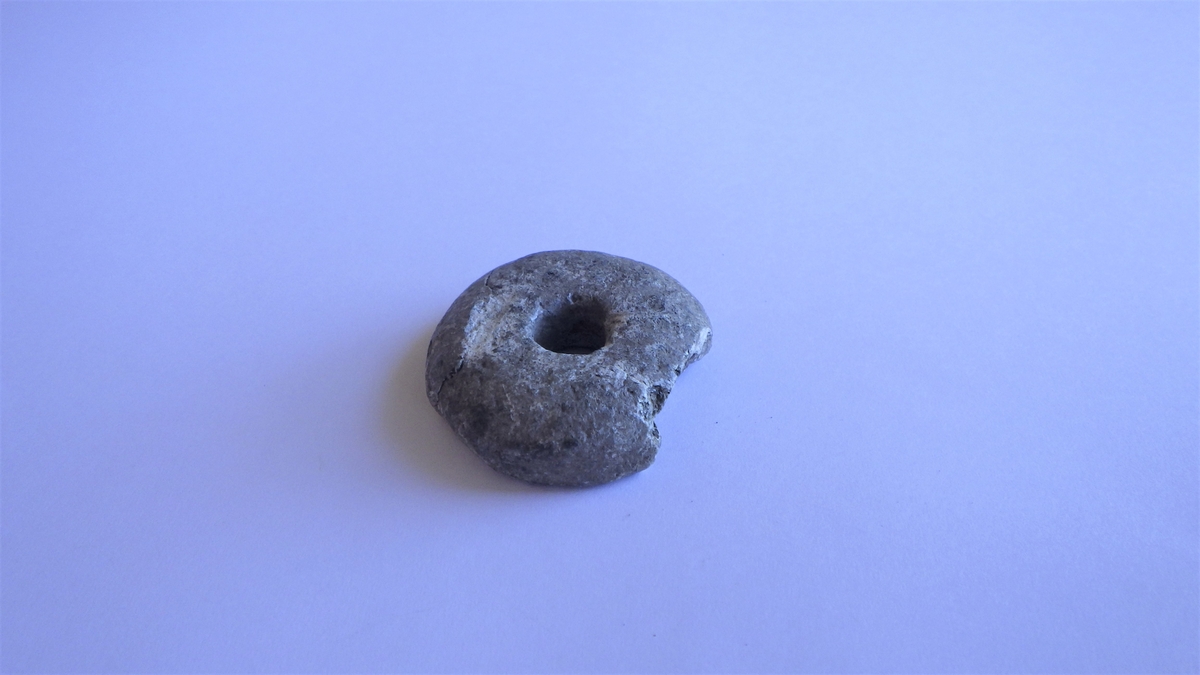 Sirkulært spinnehjul i stein, med en skade i kanten. Gjenstanden har et tilnærmet halvkuleformet utseende med flat underside og buet overside.
