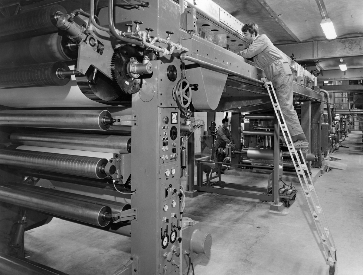 Maskinfører Roar Hovde inspiserer tørkekanalen på den nye bestryknings- og lamineringsmaskinen som ble installert ved Drammenselvens Papirfabrikker på Modum i Buskerud i 1971.  Maskinføreren var kledd i kjeledress og sto i en stige når han skulle forvisse seg om at tørkekanalen var i orden.  Den nye maskinen (BM III) ble brukt til PVDC-bestrykning (polyvinylidenklorid), samt laminere aluminiumsfolie og plastfilmer til papir og kartong, eller silikonbestryke ulike materialer.  Den var 18 meter lang, hadde en arbeidsbredde på 160 centimeter og kunne kjøres med en hastighet på opptil 200 meter i minuttet.  Maskinen var på dette tidspunktet en av de største og mest moderne i Skandinavia.  Maskinen var for øvrig montert i forlengelsen av en eldre bestryknings- og lamineringsmaskin, og de to maskinene kunne kjøres sammen, som en maskin.  På den måten kunne man, om ønskelig kjøre papir som skulle bestrykes med flere lag på hver side.  Maskinene ble brukt til å produsere papir med barrieremateriale, slik at det for eksempel kunne brukes til pakking av produkter der det var viktig å holde på aromastoffer, fuktighet, fett eller ulike kjemikalier.  Drammenselvens Papirfabrikker leverte spesialpapir til smør- og margarinpakninger, kartongbegere, iskremomslag, frysepapir og emballasje for bakeri-, konserves- og tobakksindustrien.  I tørkepartiet som ble inspisert på dette fotografiet ble bestrykningslagene herdet, mellom bestrykninger og før avsluttende opprulling. 