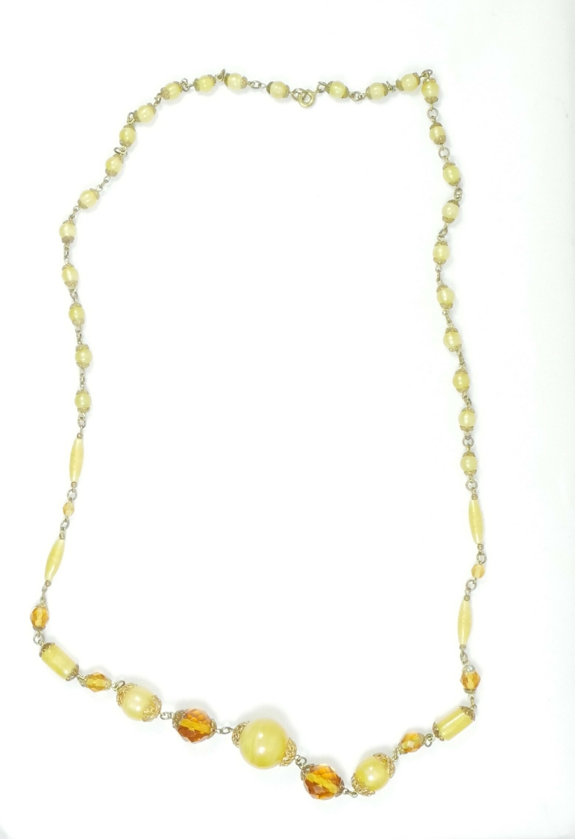 Hvit rektangulær pappemballasje med lokk. Inne i esken er det tre halskjeder. 

Halssmykke med gule steiner. 

Halssmykke to tekstiltråder med hvite steiner, en lås i enden

Gullfarget halssmykke 