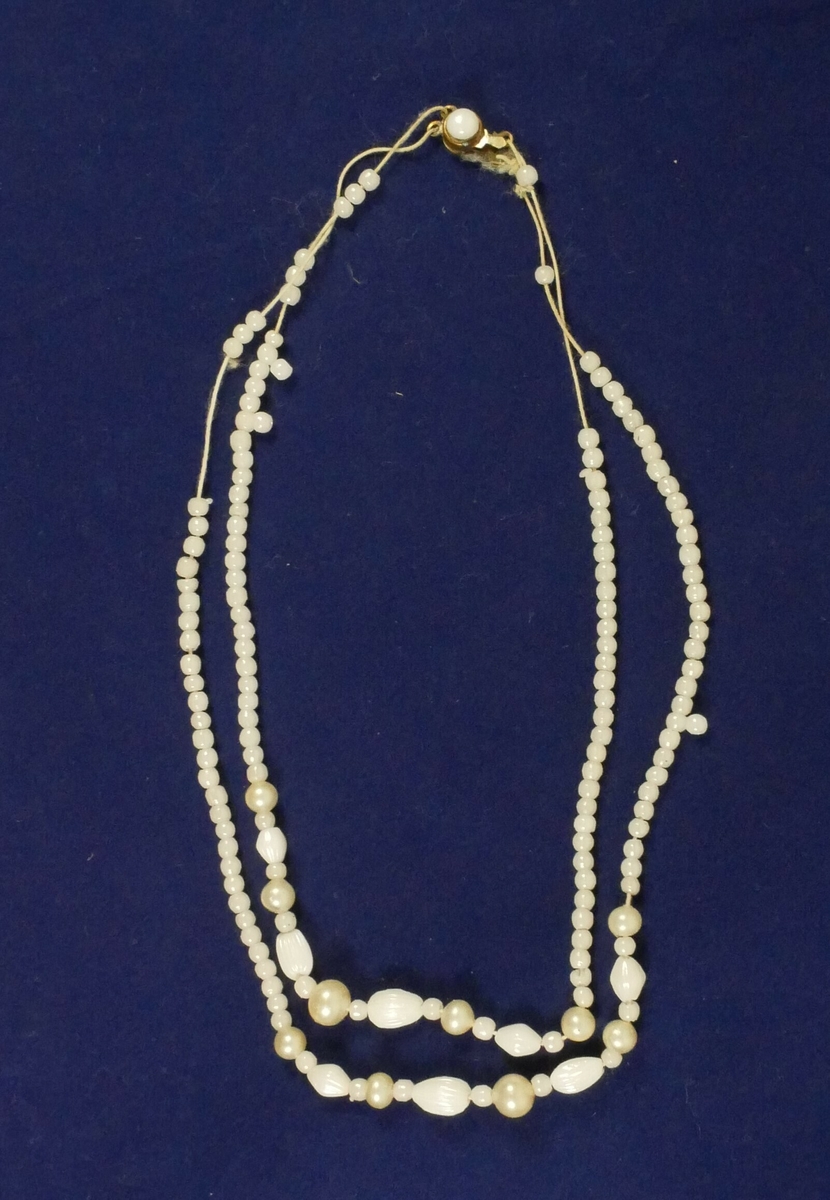 Hvit rektangulær pappemballasje med lokk. Inne i esken er det tre halskjeder. 

Halssmykke med gule steiner. 

Halssmykke to tekstiltråder med hvite steiner, en lås i enden

Gullfarget halssmykke 