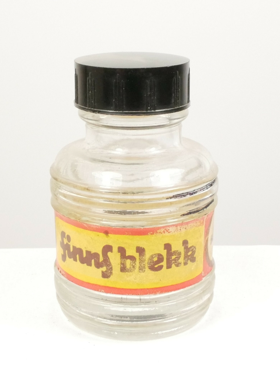 Rund klar glassflaske som smalner inn i en tut som er tettet med en svart plastkork. Rundt flasken er det en papiretikett. 