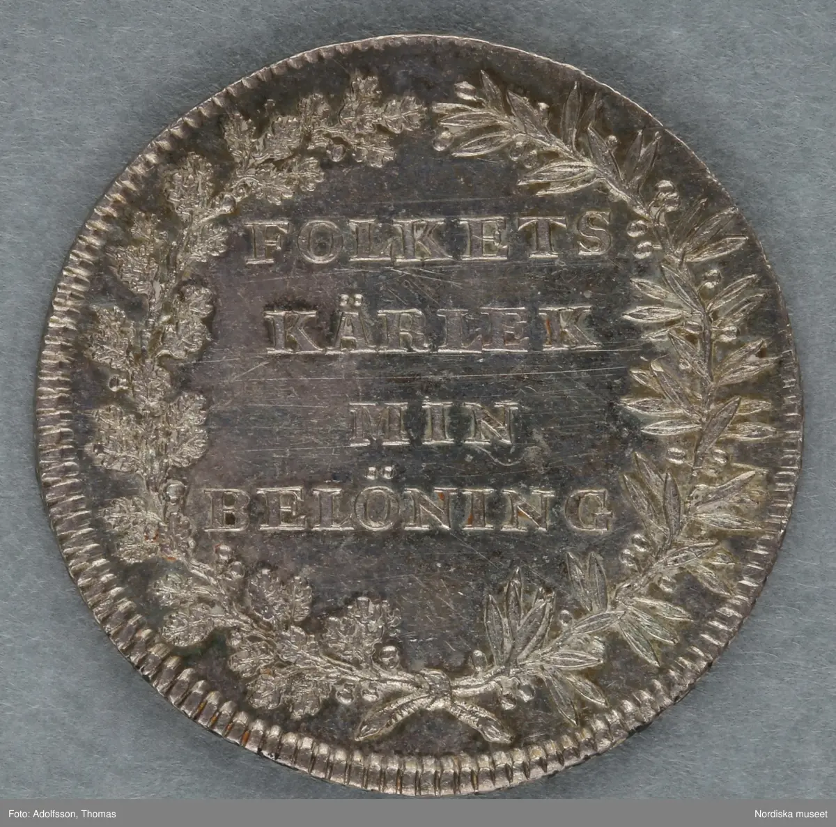 Huvudliggare:
"Minnespenningar. 3 st. I silfver. Öfver Carl XIV Johan.
a. Jfr. Hildebrand sid. 336, nr 29
b. Jfr. -"- sid.337, nr 25
c. Jfr. -"- sid 364, nr 77a"

B:
Minnespenning av silver, till minne av Carl XIV Johans kröning till kung Stockholms storkyrka den 11 maj 1818. På åtsidan kungens bröstbild och texten: "CARL XIV JOHAN SV. OCH NORR: K: KRÖNT ÅR 1818". På frånsidan kungens valspråk: "FOLKETS KÄRLEK MIN BELÖNING" inom en krans av hälften ek (symbolväxt för Norge) till hälften oliv (Sverige).
 Denna penning kastades ut bland folket på stadens torg och öppna platser vid kröningen, enligt gammalt bruk.
Carl XIV Johan var unionskung och kung över båda kungarikena Sverige och Norge.
Se Hildebrand, del 2, nr.25. Jämför inv.nr. 29 574b, som är motsvarande minnespenning från Carl XIV Johans kröning i Trondheims domkyrka samma år.
Maria Maxén/2005