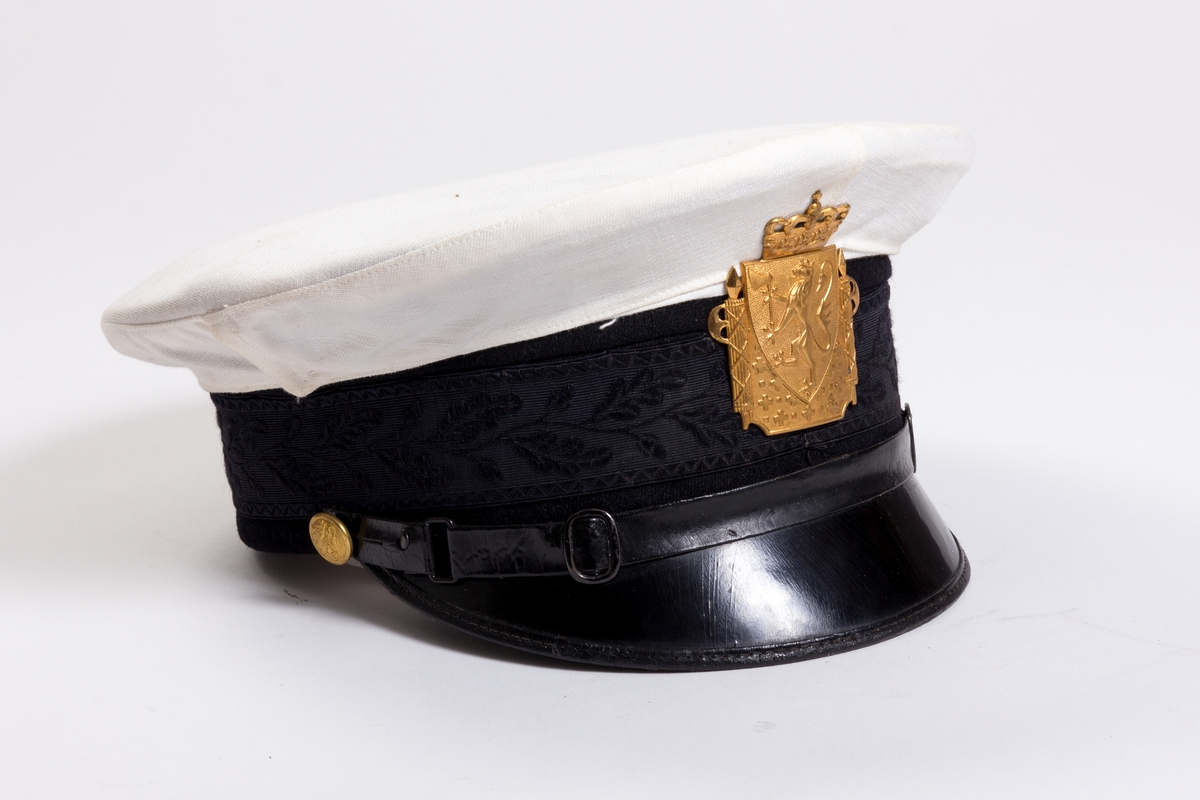 Uniformslue med hvitt avtagbart trekk og med sort brem og luering. Gullfarget rektangulært luemerke med motiv av den norske løve med krone. 
Luering med svettereim i brunt kunstlær og trukket i et sort silke?stoff i vevd bladmønster. Lærreim over skjerm er festet med en gullknapp på hver side.