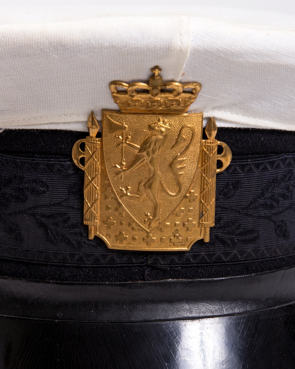 Uniformslue med hvitt avtagbart trekk og med sort brem og luering. Gullfarget rektangulært luemerke med motiv av den norske løve med krone. 
Luering med svettereim i brunt kunstlær og trukket i et sort silke?stoff i vevd bladmønster. Lærreim over skjerm er festet med en gullknapp på hver side.