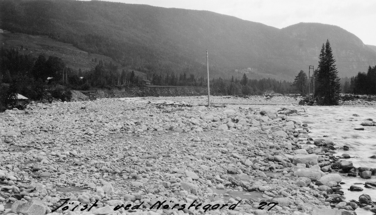 Elvelandskap i Gøyst, et vassdrag i Tinn i Telemark.  Fotografiet ble tatt i 1927, i nærheten av Nørstegard, ovenfor Ullern bru.  Disse stedene ligger ved den nedre delen av Gøyst, der dalføret er forholdsvis flatbotnet og vidt.  Vi ser da også at elveløpet er forholdsvis vidt, men at det på opptakstidspunktet bare gikk vann i djupålen til høyre i bildet.  Til venstre i bildet har flomvann skylt med seg humusjorda, slik at det som er blitt liggende igjen er ei stein- og grusør, der fløtingstømmeret antakelig hadde lett for å sette seg fast.  Da dette fotografiet ble tatt var imidlertid tømmeret utfløtet.  1927 var et flomår da vannmassene i elevene i Skiensvassdraget gjorde betydelig skade på tilstøtende åker- og engarealer mange steder.  I rapporten dette fotografiet er hentet fra heter det om Gøyst at «… Flommene er ofte voldsomme og kommer meget hurtig.  Den har derfor foraarsaket betydelige elvebrudd og tilsvarende avleiringer, spesielt i 1. rote, hvor grunnen er utelukkende aur og sand.  I storflommen i 1927 tok elven igjen det leie den forlot i flommen 1879 og voldte betydelig skade på eiendommene i 1. rote.»  Antakelig er det dette fotografen har forsøkt å dokumentere med dette fotografiet.  Gøyst var fløtbar fra det stedet der Nysetåa og Gjerdøla løper sammen, cirka 10 kilometer fra elveosen i nordenden av Tinnsjøen.  På denne strekningen var det et samlet fall på cirka 260 meter.  Fløtinga her var fordelt på tre roter, og den sysselsatte cirka ti mann i sesongen.