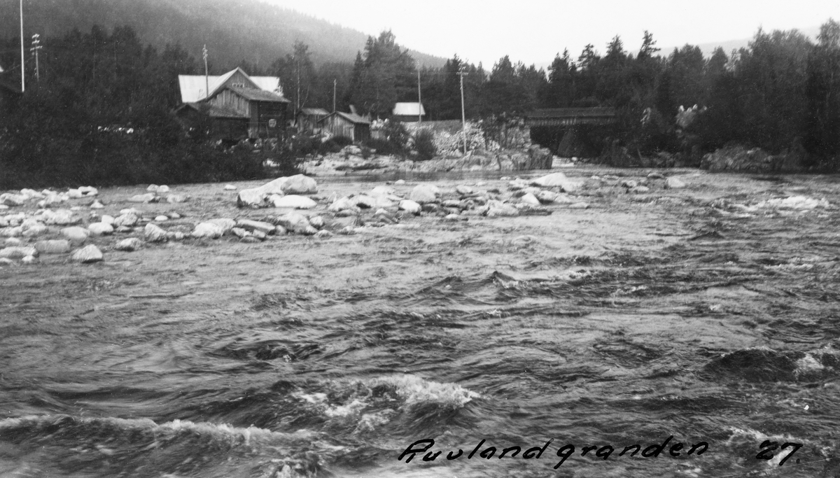 Nedre del av elva Austbygdåe i Tinn i Telemark.  I går elva, forholdsvis flomstor, i en slak kurve med en steinrygg på den ene sida, en såkalt «grande».  Akkurat denne ble kalt «Ruvlandgranden» etter garden elva passerte på dette stedet, antakelig den eiendommen vi ser bebyggelsen på til venstre i bildeflata.  Dalføret omkring var forholdsvis vidt, og i stor utstrekning skogbevokst.  Austbygdåe kom fra Tessungsjø, og distansen derfra til utlåpet ved Tosand i nordenden av Tinnsjøen var på drøyt 30 kilometer.  Ettersom Tessungsjå ligger om lag 1 400 meter over havet, var det lite tømmer å fløte ved den øverste delen av Austbygdåe.  Enkelte år ble det fløtet fra Kongsbergsætra, snaut 900 meter over havet.  Austbygdåe hadde sju fløtingsroter, men på grunn av høytliggende terreng og forholdsvis lite tømmerskog, var det slett ikke hvert år det ble levert tømmer i de to-tre øverste rotene.  Ruvlandgranden lå altså i den nedre delen av vassdraget, der grensa mellom første og andre rode gikk ved brua vi skimter til høyre for gardstunet på bildet.  Fløtinga i Austbygdåe gav sesongbeskjeftigelse til 14-15 mann.  Topografien var slik at flommene kunne komme ganske brått, men alt i alt ble dette vassdraget ansett for å være forholdsvis godt å «bryte» i.