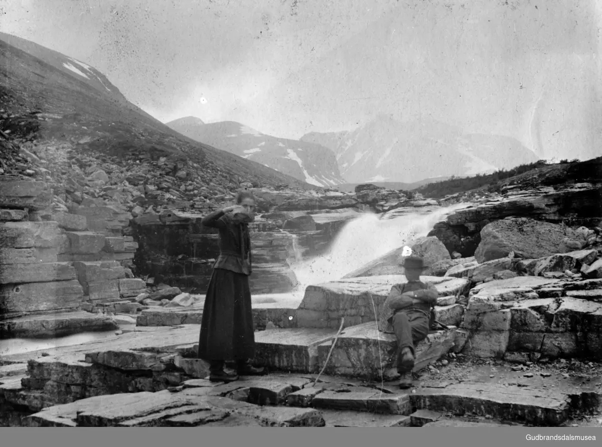 To turistar ved ein foss i Langglupdalen. Rondeslottet i bakgrunnen.