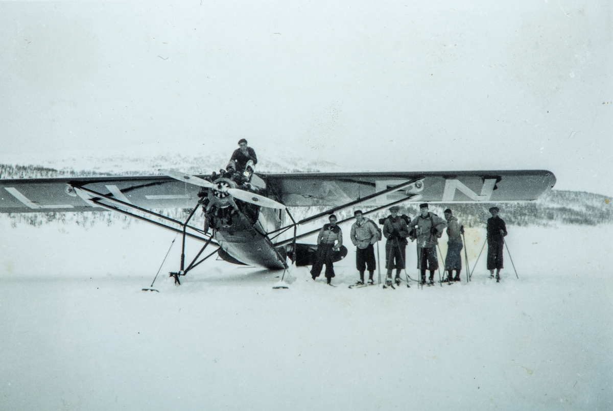 Gruppe 7 ukjente, 1 fly på bakken. Snø,Vinter. Stinson SM-1, LN-ABN fra Norsk Lufttrafikk.
1 mann sittende på flyet, resterende står med ski og sekker.
Bilde er hentet fra et album med primært bilder fra Stange området.