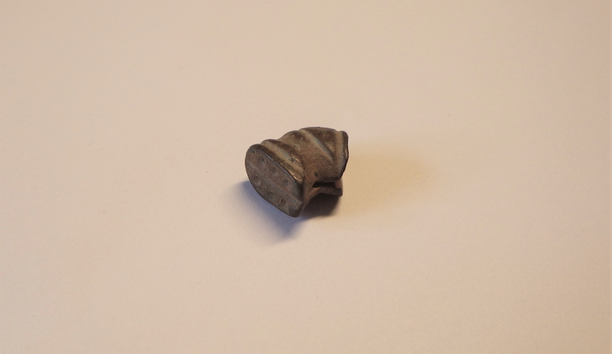 Avlang og buet metalldel med ovalt tilsnitt. Den ene enden har en snittflate som har en spalte i midten. I motsatt ende sees en glatt overflate med seks punslinger fordelt på tre stykker langs hver side av midtlinjen.  Overflaten ellers er dekorert med linjer/sirkler rundt gjenstanden. Dette kan være en knivtopp.