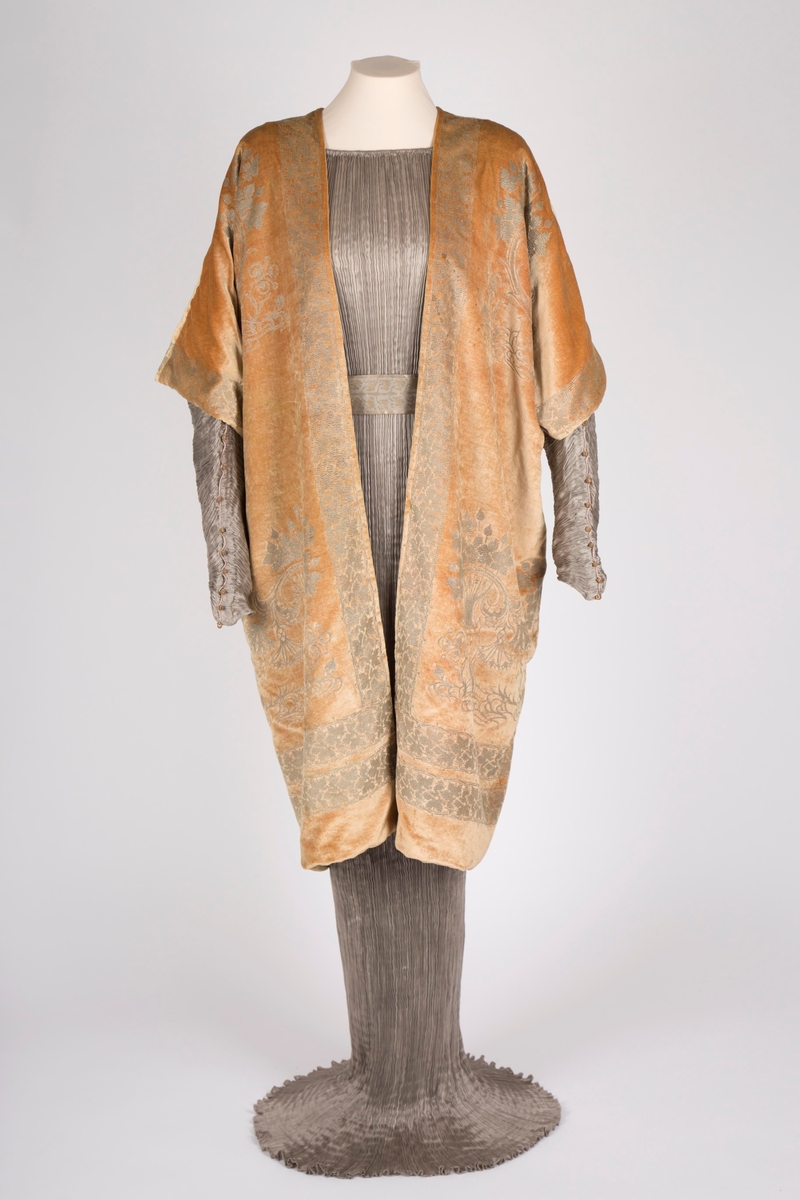 Knelang jakke av tynn, myk, aprikosfarget silkefløyel, med påtrykt mønter i sølv. Helfôret med grå, ripsartet silke. Jakken er åpen foran, med rettvinklede ermer, og er sterkt kimonoinspirert. Ermene er albulange og skåret iett med henholdsvis forstykkene og ryggstykket. For- og bakstykker er sammensydd i sidene, på skulderen og langs ermenes overside. Ryggen er helskåret, med relativ dyp V-ringning i nakken. Jakken er rikt dekorert med sølvfarget trykk av vridde treformer, blader og blomster. Hovedmotivet er plassert på hver hofte foran, og gjentatt på brystpartiet. På ryggen er dette tilsvarende plassert, det hele innrammet av en bord med stiliserte vinranker. Borden er dobbel i nederkant, men den øverste av disse bordene føres parallelt opp langs ryggens midte, fordeler seg i nakken og føres ned langs åpningen midt foran.