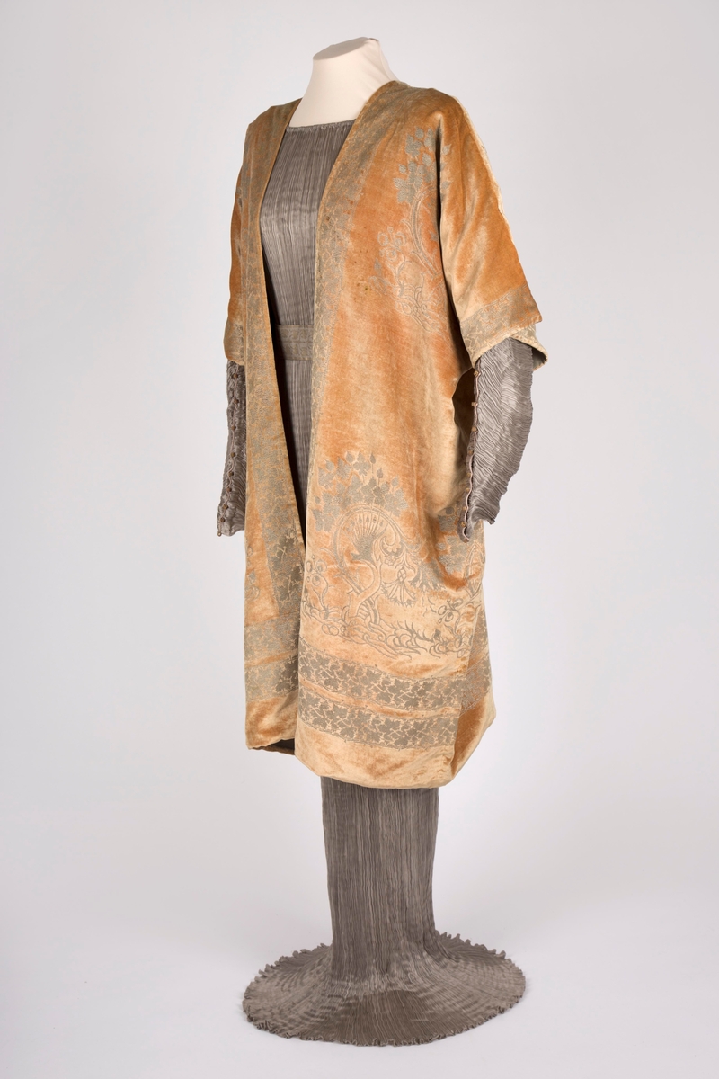 Knelang jakke av tynn, myk, aprikosfarget silkefløyel, med påtrykt mønter i sølv. Helfôret med grå, ripsartet silke. Jakken er åpen foran, med rettvinklede ermer, og er sterkt kimonoinspirert. Ermene er albulange og skåret iett med henholdsvis forstykkene og ryggstykket. For- og bakstykker er sammensydd i sidene, på skulderen og langs ermenes overside. Ryggen er helskåret, med relativ dyp V-ringning i nakken. Jakken er rikt dekorert med sølvfarget trykk av vridde treformer, blader og blomster. Hovedmotivet er plassert på hver hofte foran, og gjentatt på brystpartiet. På ryggen er dette tilsvarende plassert, det hele innrammet av en bord med stiliserte vinranker. Borden er dobbel i nederkant, men den øverste av disse bordene føres parallelt opp langs ryggens midte, fordeler seg i nakken og føres ned langs åpningen midt foran.