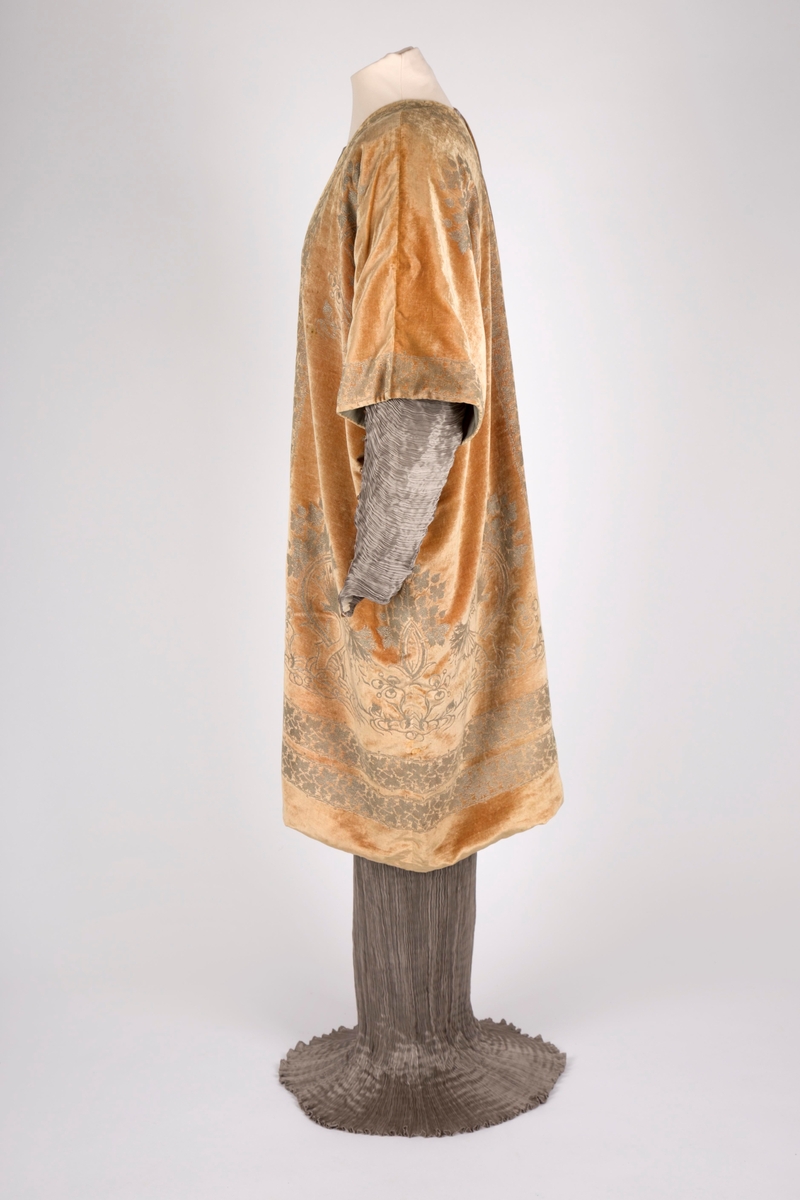 Knelang jakke av tynn, myk, aprikosfarget silkefløyel, med påtrykt mønter i sølv. Helfôret med grå, ripsartet silke. Jakken er åpen foran, med rettvinklede ermer, og er sterkt kimonoinspirert. Ermene er albulange og skåret iett med henholdsvis forstykkene og ryggstykket. For- og bakstykker er sammensydd i sidene, på skulderen og langs ermenes overside. Ryggen er helskåret, med relativ dyp V-ringning i nakken. Jakken er rikt dekorert med sølvfarget trykk av vridde treformer, blader og blomster. Hovedmotivet er plassert på hver hofte foran, og gjentatt på brystpartiet. På ryggen er dette tilsvarende plassert, det hele innrammet av en bord med stiliserte vinranker. Borden er dobbel i nederkant, men den øverste av disse bordene føres parallelt opp langs ryggens midte, fordeler seg i nakken og føres ned langs åpningen midt foran.