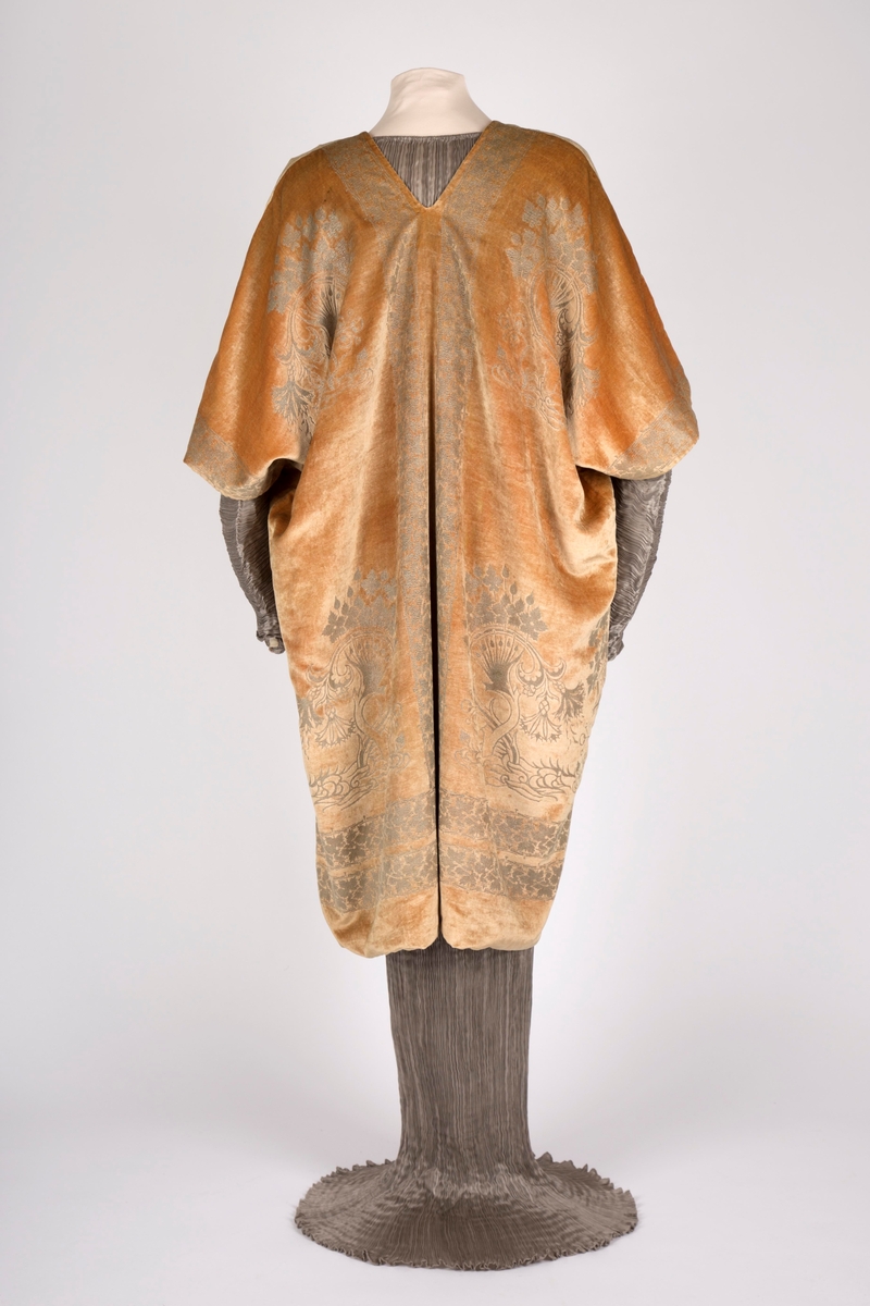 Knelang jakke av tynn, myk, aprikosfarget silkefløyel, med påtrykt mønter i sølv. Helfôret med grå, ripsartet silke. Jakken er åpen foran, med rettvinklede ermer, og er sterkt kimonoinspirert. Ermene er albulange og skåret iett med henholdsvis forstykkene og ryggstykket. For- og bakstykker er sammensydd i sidene, på skulderen og langs ermenes overside. Ryggen er helskåret, med relativ dyp V-ringning i nakken. Jakken er rikt dekorert med sølvfarget trykk av vridde treformer, blader og blomster. Hovedmotivet er plassert på hver hofte foran, og gjentatt på brystpartiet. På ryggen er dette tilsvarende plassert, det hele innrammet av en bord med stiliserte vinranker. Borden er dobbel i nederkant, men den øverste av disse bordene føres parallelt opp langs ryggens midte, fordeler seg i nakken og føres ned langs åpningen midt foran.