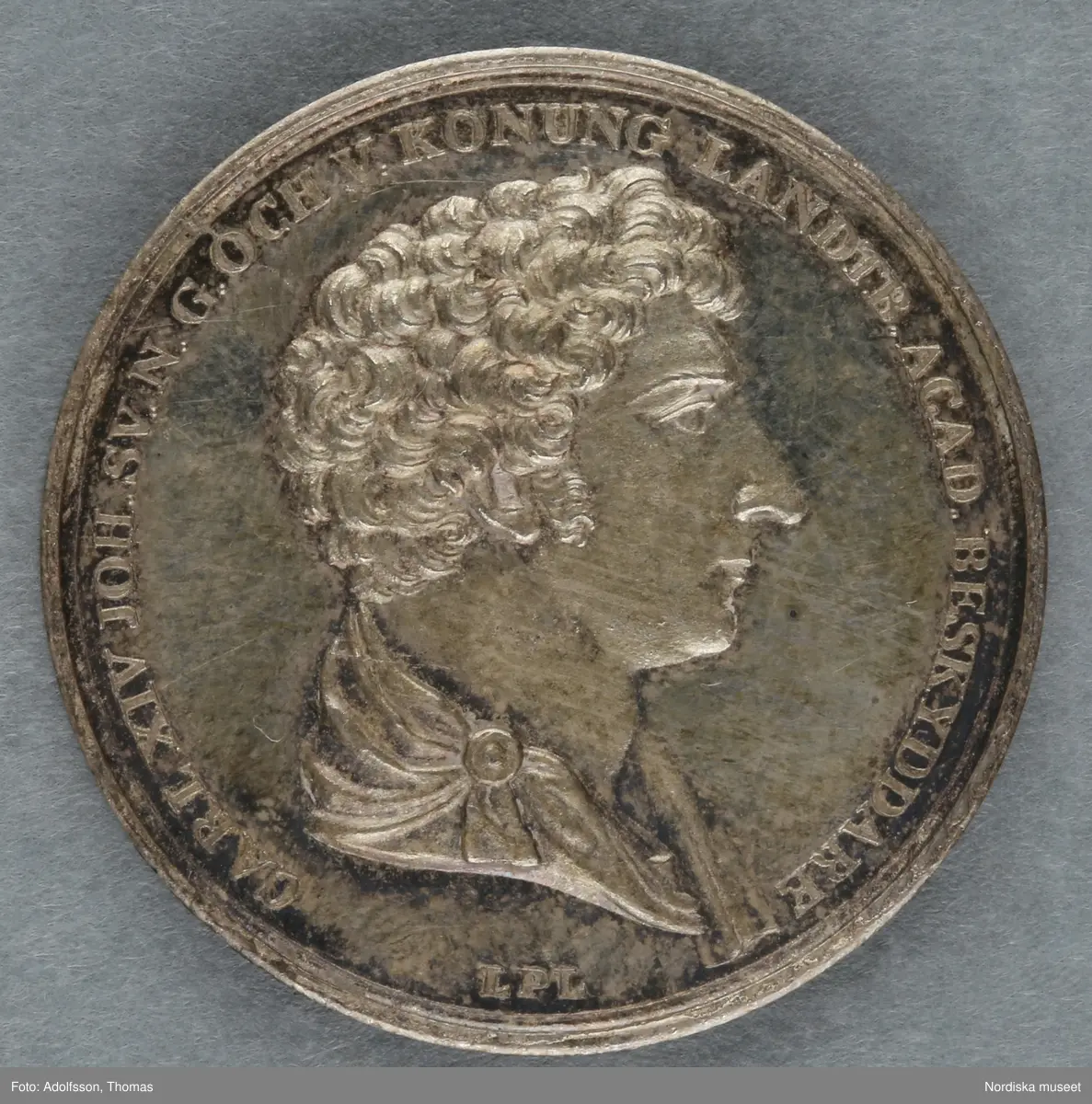 Huvudliggare:
"Minnespenningar. 3 st. I silfver. Öfver Carl XIV Johan.
a. Jfr. Hildebrand sid. 336, nr 29
b. Jfr. -"- sid.337, nr 25
c. Jfr. -"- sid 364, nr 77a"

B:
Minnespenning av silver, till minne av Carl XIV Johans kröning till kung Stockholms storkyrka den 11 maj 1818. På åtsidan kungens bröstbild och texten: "CARL XIV JOHAN SV. OCH NORR: K: KRÖNT ÅR 1818". På frånsidan kungens valspråk: "FOLKETS KÄRLEK MIN BELÖNING" inom en krans av hälften ek (symbolväxt för Norge) till hälften oliv (Sverige).
 Denna penning kastades ut bland folket på stadens torg och öppna platser vid kröningen, enligt gammalt bruk.
Carl XIV Johan var unionskung och kung över båda kungarikena Sverige och Norge.
Se Hildebrand, del 2, nr.25. Jämför inv.nr. 29 574b, som är motsvarande minnespenning från Carl XIV Johans kröning i Trondheims domkyrka samma år.
Maria Maxén/2005