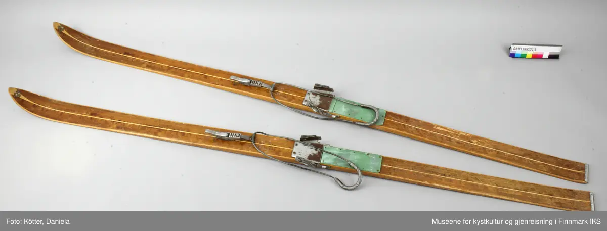 Tofarget treski med Kandaharbindinger. Skiene er mørke og har en lys stripe i midten. Helt foran på spissene er det påført firmalogoen til Madshus.  Bak bindingen er det spikret på biter av grønn linoleum eller kunststoff. Helt bak har skiene en metallkant. Foran bindingene er det slagmerker med tallene 210 og 567. 
