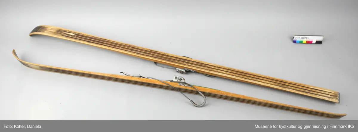 Tofarget treski med Kandaharbindinger. Skiene er mørke og har en lys stripe i midten. Helt foran på spissene er det påført firmalogoen til Madshus.  Bak bindingen er det spikret på biter av grønn linoleum eller kunststoff. Helt bak har skiene en metallkant. Foran bindingene er det slagmerker med tallene 210 og 567. 
