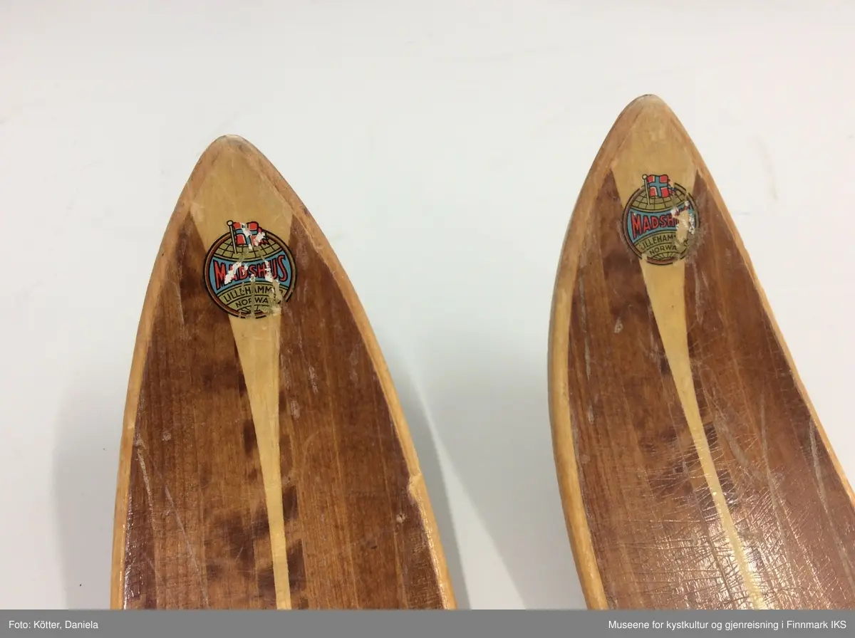 Tofarget treski med Kandaharbindinger. Skiene er mørke og har en lys stripe i midten. Helt foran på spissene er det påført firmalogoen til Madshus.  Bak bindingen er det spikret på biter av grønn linoleum eller kunststoff. Helt bak har skiene en metallkant. Foran bindingene er det slagmerker med tallene 210 og 567. 
