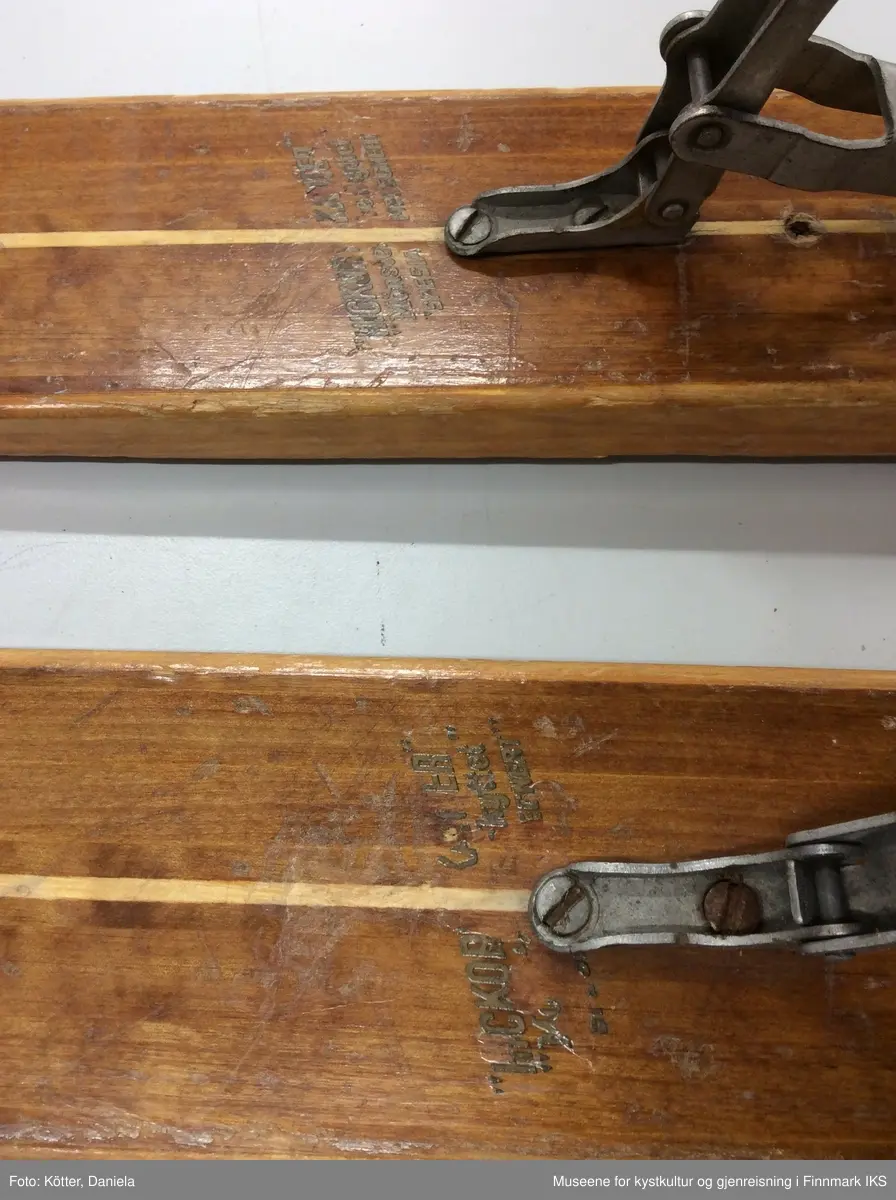 Tofarget treski med Kandaharbindinger. Skiene er mørke og har en lys stripe i midten. Helt foran på spissene er det påført firmalogoen til Madshus.  Bak bindingen er det spikret på biter av grønn linoleum eller kunststoff. Helt bak har skiene en metallkant. Foran bindingene er det slagmerker med tallene 210 og 567. 
