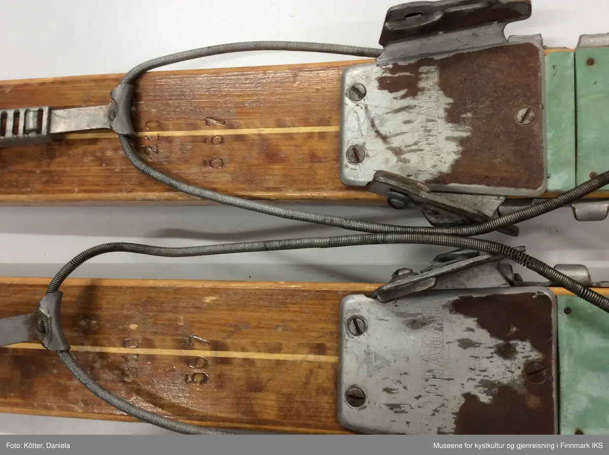 Tofarget treski med Kandaharbindinger. Skiene er mørke og har en lys stripe i midten. Helt foran på spissene er det påført firmalogoen til Madshus.  Bak bindingen er det spikret på biter av grønn linoleum eller kunststoff. Helt bak har skiene en metallkant. Foran bindingene er det slagmerker med tallene 210 og 567. 
