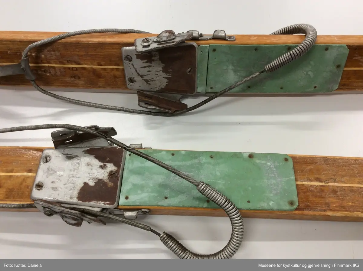 Tofarget treski med Kandaharbindinger. Skiene er mørke og har en lys stripe i midten. Helt foran på spissene er det påført firmalogoen til Madshus.  Bak bindingen er det spikret på biter av grønn linoleum eller kunststoff. Helt bak har skiene en metallkant. Foran bindingene er det slagmerker med tallene 210 og 567. 
