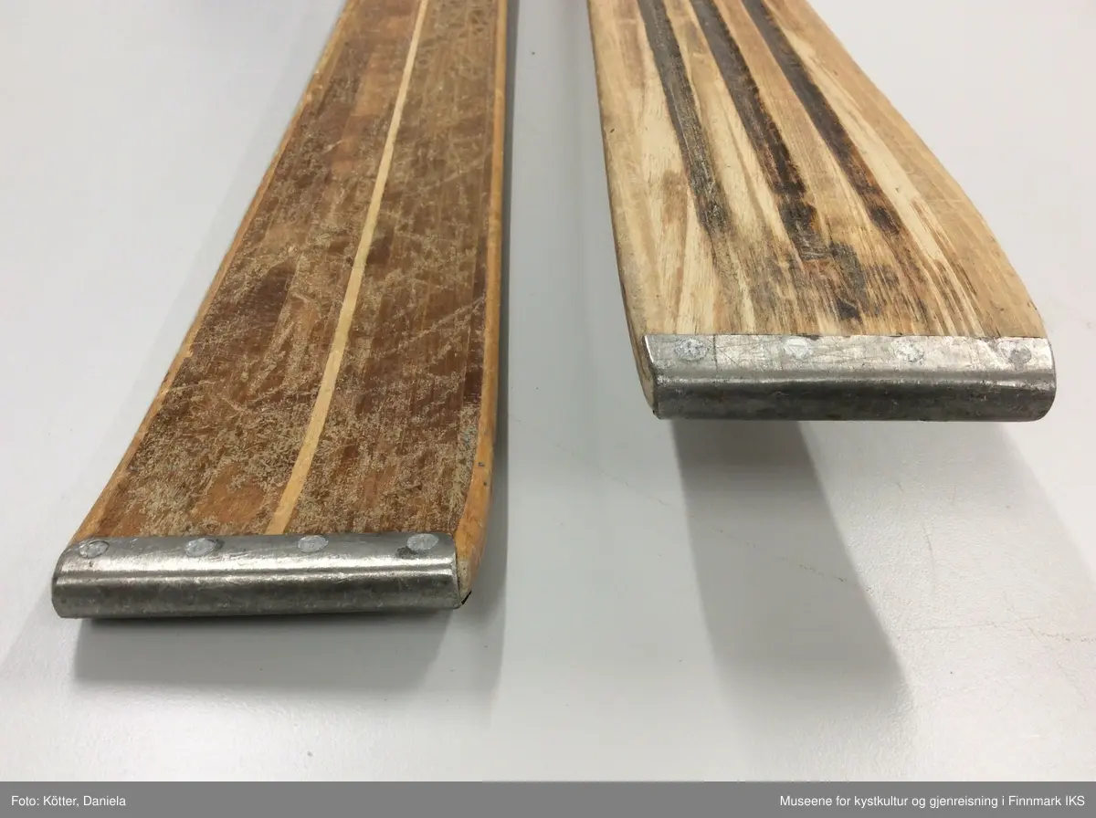 Tofarget treski med Kandaharbindinger. Skiene er mørke og har en lys stripe i midten. Helt foran på spissene er det påført firmalogoen til Madshus.  Bak bindingen er det spikret på biter av grønn linoleum eller kunststoff. Helt bak har skiene en metallkant. Foran bindingene er det slagmerker med tallene 210 og 567. 
