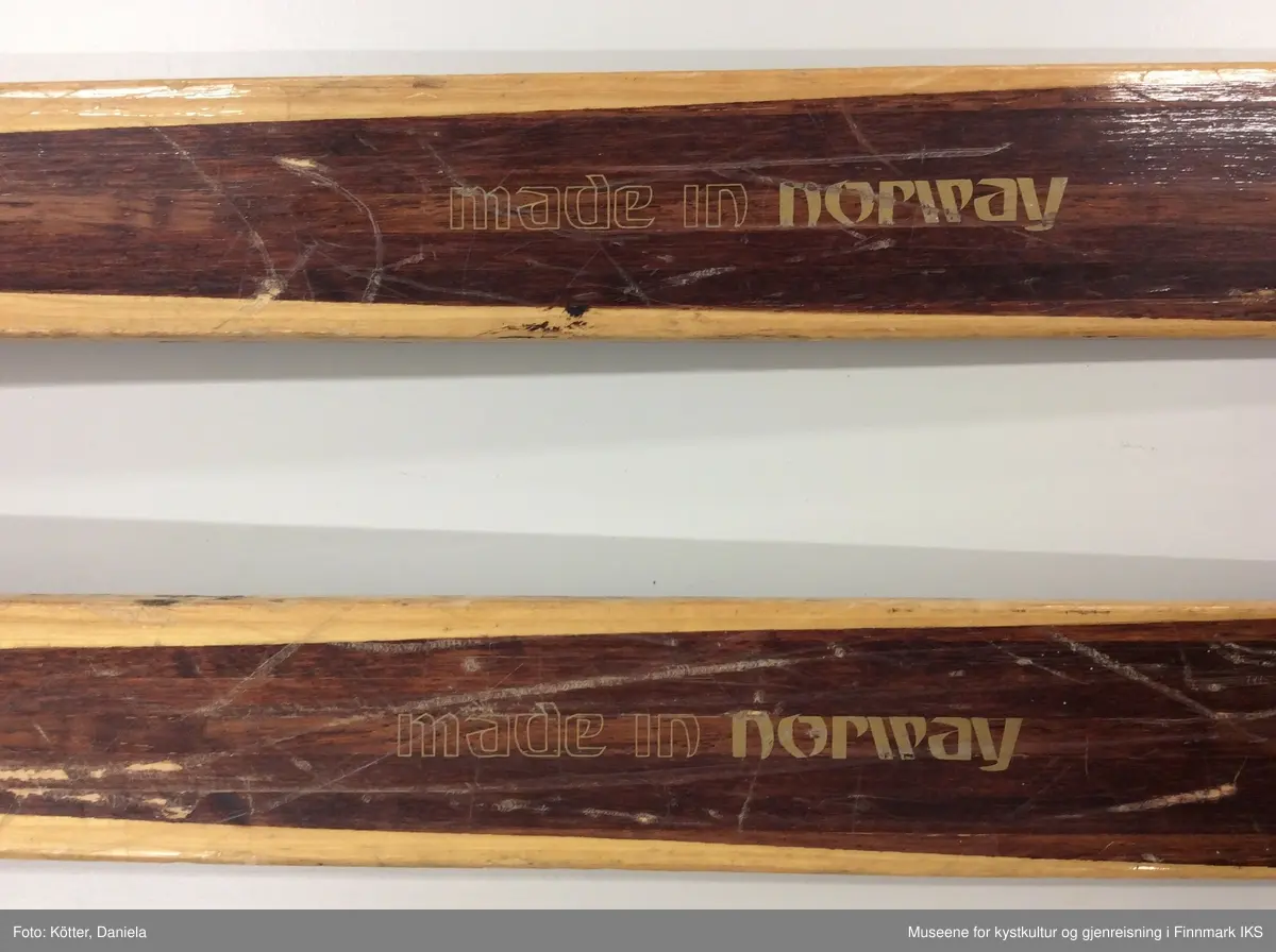 Tofarget treski med Kandaharbindinger. Bak bindingen er det påført deler i rød kunststoff for bedre feste med skoen. Helt bak har skiene en metallkant.