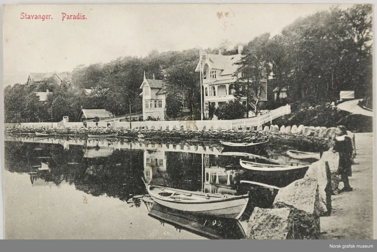 Postkort med motiv fra Paradis, innerst ved Hillevågsvatnet i Stavanger. To jenter står på en vei og ser utover vannet. Robåter ligger fortøyd til land