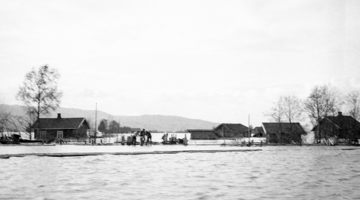 Storflom ved Fetsund våren 1916.  Fotografiet viser ei elveslette med flere små enetasjes stuebygninger med tilhørende uthusbygninger og omliggende jordbruksareal.  Sletta er oversvømt av flomvann slik at bygningene står i vann.  På vannflata i forgrunnen ligger det en føytende, lenseliknende konstruksjon Bakenfor, ved et gjerde, skimtes noen karer som antakelig arbeider med å berge et eller annet. 