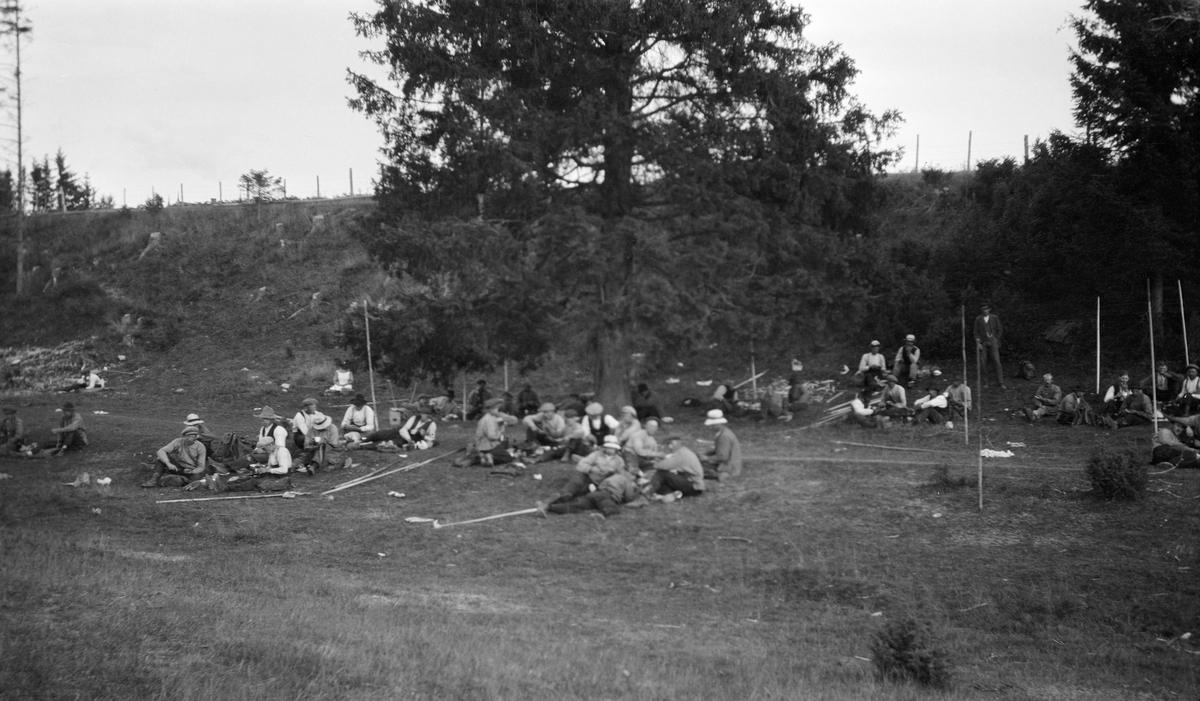 Kvilestund for mannskaper ved attholdeslensa ved Bingen i Sørum, Akershus, sommeren 1917.  Fotografen har antakelig stått med ryggen mot elva og tatt bildet mot den grasbevokste strandbakken, der en god del mannfolk sitter og ligger på bakken og kviler seg omkring ei gran. En del av dem har fløterhaker liggende på bakken, andre er stauret ned i torva.  På bakkekammen i bakgrunnen skimtes et trådgjerde, antakelig en avgrensing av et jordbruksareal. 

Det året dette fotografiet ble tatt var det anmeldt 12 308 986 tømmerstokker til fløting i Glommavassdraget ovenfor Fetsund.  Dette tømmeret hadde et samlet volum på 2 169 521 kubikkmeter. 

En kort historikk om Bingen lenser finnes under fanen «Opplysninger». 