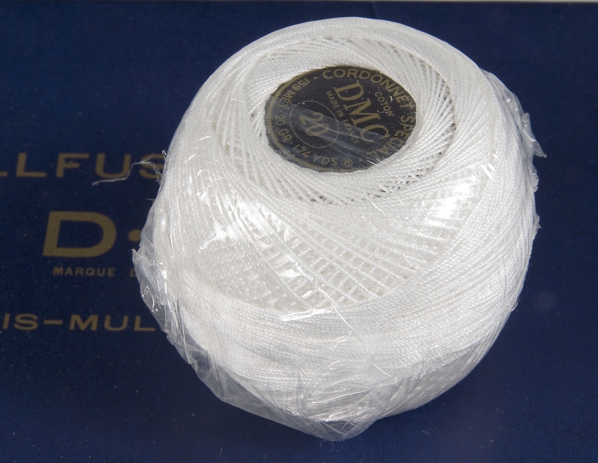 Ett inplastat nystan av vit bomullstråd, troligen virkgarn. Svart etikett med guldfärgad tryckt text: CORDONNET SPÉCIAL COTON DMC, MADE IN FRANCE. Låg i ask UM26115a.