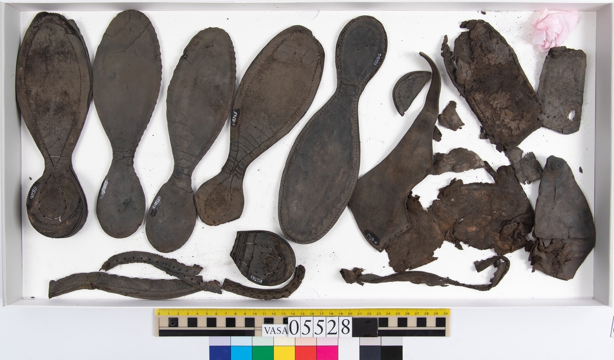 Dokumentation 1997:
Ett tjugotal delar och fragment från ett par skor.
Del av en skobotten, fyndnummer 05528. Skobottnen består av två sulor mellan vilka klacklappar sitter fästade. Sulorna och klacklapparna har monterats samman med tråd av konserveringsenheten.
En klack, fyndnummer 05528:1. Klacken är uppbyggd av sju klacklappar. Klacken har monterats samman med tråd av konserveringsenheten.
Två sulor, fyndnummer 05528:2 samt 05528:3. Sula fyndnummer 05528:3 har ett nedslitet hälparti. Sulorna och klacken är delar från den andra skobottnen som bildar par med skobotten fyndnummer 05528.
Två sulor, fyndnummer 05528:4 och 05528:5. Sulorna hör samman med någon av de två ovan nämnda skobottnarna.
Ett sidosycke med slejf, fyndnummer 05528:6. Längs med sidostyckets nedre kant löper stygnshål.
Ett 15-tal delar och fragment av holtrand, sulor mm.
Lädret är kraftigt och välbevarat.

Dokumentation 2022:
Fragment från ovanläder och ett par skobottnar (05528:4). Framstycke och sulor till öppna randskor. Tillverkade i läder. Skapades på en rak läst och bars på höger fot. Det finns regelbundna skurna dekorationer på framstycket. Klacken är reparerad med en läderlapp under sulan. Det finns tecken på delaminering på framstycket. Det finns hål i skobottens sidor och i mittensulan. Det finns inga tecken på lagningar.
För mer information, se Uppritning under Länkade filer.

Text in english:
These are one vamp fragment and a pair of bottom units (05528:4) of open side latchet shoes, made of leather, of randed construction with a patch heel, made on a straight last and worn on the right foot, with decorative pinked features across the vamp. The vamps are delaminated, and the bottom unit presents holes on both lateral sides and on the midsole. There are no signs of repairs.
For more information, go to Länkade filer: Uppritning