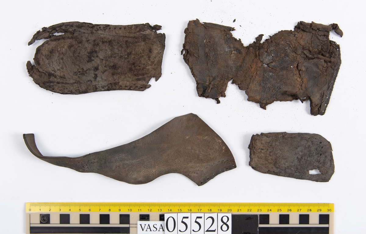 Dokumentation 1997:
Ett tjugotal delar och fragment från ett par skor.
Del av en skobotten, fyndnummer 05528. Skobottnen består av två sulor mellan vilka klacklappar sitter fästade. Sulorna och klacklapparna har monterats samman med tråd av konserveringsenheten.
En klack, fyndnummer 05528:1. Klacken är uppbyggd av sju klacklappar. Klacken har monterats samman med tråd av konserveringsenheten.
Två sulor, fyndnummer 05528:2 samt 05528:3. Sula fyndnummer 05528:3 har ett nedslitet hälparti. Sulorna och klacken är delar från den andra skobottnen som bildar par med skobotten fyndnummer 05528.
Två sulor, fyndnummer 05528:4 och 05528:5. Sulorna hör samman med någon av de två ovan nämnda skobottnarna.
Ett sidosycke med slejf, fyndnummer 05528:6. Längs med sidostyckets nedre kant löper stygnshål.
Ett 15-tal delar och fragment av holtrand, sulor mm.
Lädret är kraftigt och välbevarat.

Dokumentation 2022:
Fragment från ovanläder och ett par skobottnar (05528:4). Framstycke och sulor till öppna randskor. Tillverkade i läder. Skapades på en rak läst och bars på höger fot. Det finns regelbundna skurna dekorationer på framstycket. Klacken är reparerad med en läderlapp under sulan. Det finns tecken på delaminering på framstycket. Det finns hål i skobottens sidor och i mittensulan. Det finns inga tecken på lagningar.
För mer information, se Uppritning under Länkade filer.

Text in english:
These are one vamp fragment and a pair of bottom units (05528:4) of open side latchet shoes, made of leather, of randed construction with a patch heel, made on a straight last and worn on the right foot, with decorative pinked features across the vamp. The vamps are delaminated, and the bottom unit presents holes on both lateral sides and on the midsole. There are no signs of repairs.
For more information, go to Länkade filer: Uppritning