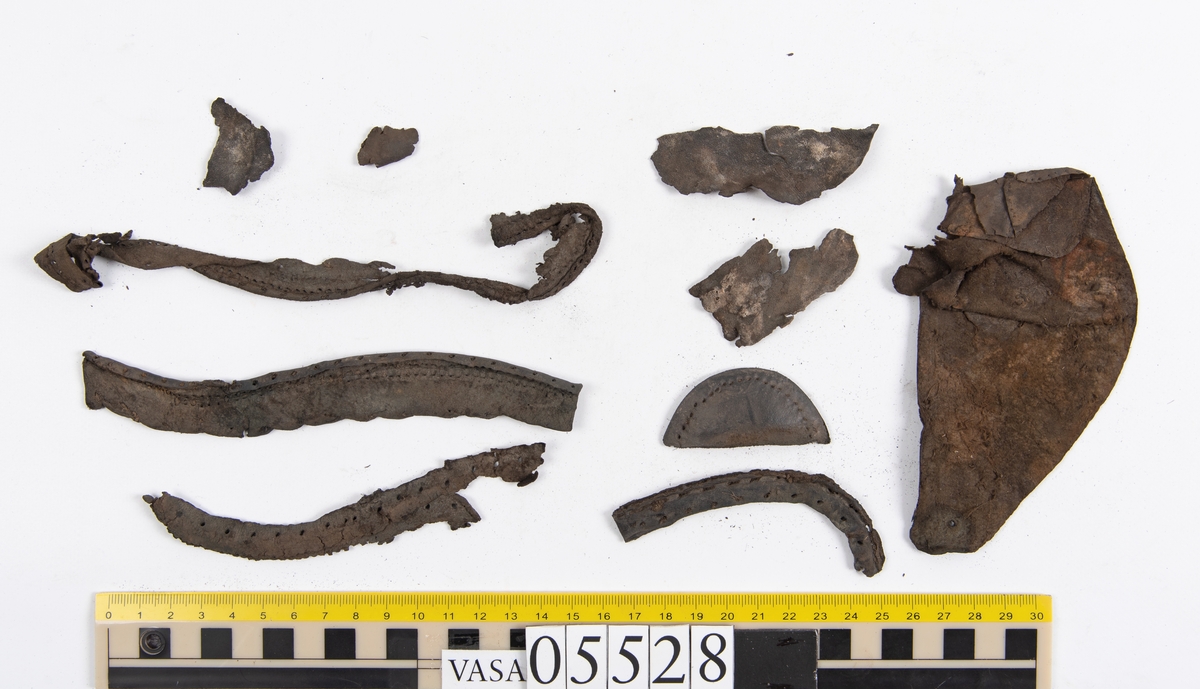 Dokumentation 1997:
Ett tjugotal delar och fragment från ett par skor.
Del av en skobotten, fyndnummer 05528. Skobottnen består av två sulor mellan vilka klacklappar sitter fästade. Sulorna och klacklapparna har monterats samman med tråd av konserveringsenheten.
En klack, fyndnummer 05528:1. Klacken är uppbyggd av sju klacklappar. Klacken har monterats samman med tråd av konserveringsenheten.
Två sulor, fyndnummer 05528:2 samt 05528:3. Sula fyndnummer 05528:3 har ett nedslitet hälparti. Sulorna och klacken är delar från den andra skobottnen som bildar par med skobotten fyndnummer 05528.
Två sulor, fyndnummer 05528:4 och 05528:5. Sulorna hör samman med någon av de två ovan nämnda skobottnarna.
Ett sidosycke med slejf, fyndnummer 05528:6. Längs med sidostyckets nedre kant löper stygnshål.
Ett 15-tal delar och fragment av holtrand, sulor mm.
Lädret är kraftigt och välbevarat.

Dokumentation 2022:
Fragment från ovanläder och ett par skobottnar (05528:4). Framstycke och sulor till öppna randskor. Tillverkade i läder. Skapades på en rak läst och bars på höger fot. Det finns regelbundna skurna dekorationer på framstycket. Klacken är reparerad med en läderlapp under sulan. Det finns tecken på delaminering på framstycket. Det finns hål i skobottens sidor och i mittensulan. Det finns inga tecken på lagningar.
För mer information, se Uppritning under Länkade filer.

Text in english:
These are one vamp fragment and a pair of bottom units (05528:4) of open side latchet shoes, made of leather, of randed construction with a patch heel, made on a straight last and worn on the right foot, with decorative pinked features across the vamp. The vamps are delaminated, and the bottom unit presents holes on both lateral sides and on the midsole. There are no signs of repairs.
For more information, go to Länkade filer: Uppritning