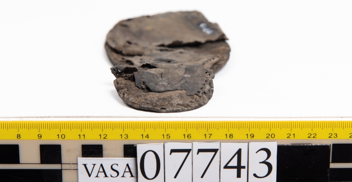 En sula. Sulan ligger vikt. Kvarsittande tråd samt hål efter skopligg. Lädret är spaltat. Två lösa mindre fragment.