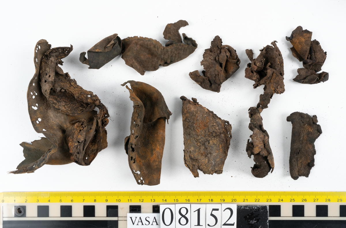 Ett femtiotal större och mindre delar och fragment från två stövlar: sulor, inlägg, ovanläder mm. Lädret ligger vikt. Bland delarna finns också en större klump av tråd eller snöre. Även ulltrådar.

Lädret är hårt och sprött samt har gul-vita utfällningar.