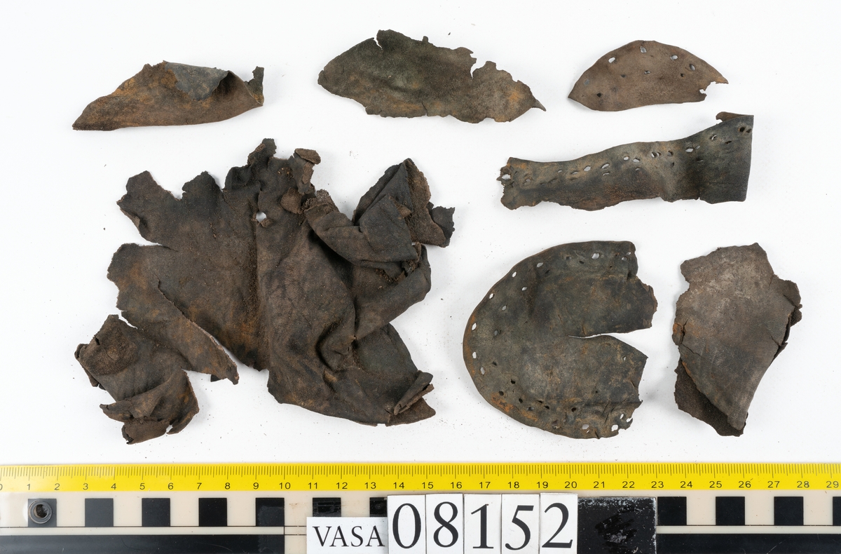Ett femtiotal större och mindre delar och fragment från två stövlar: sulor, inlägg, ovanläder mm. Lädret ligger vikt. Bland delarna finns också en större klump av tråd eller snöre. Även ulltrådar.

Lädret är hårt och sprött samt har gul-vita utfällningar.