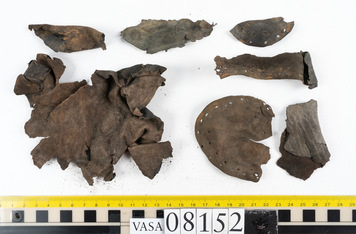 Ett femtiotal större och mindre delar och fragment från två stövlar: sulor, inlägg, ovanläder mm. Lädret ligger vikt. Bland delarna finns också en större klump av tråd eller snöre. Även ulltrådar.

Lädret är hårt och sprött samt har gul-vita utfällningar.