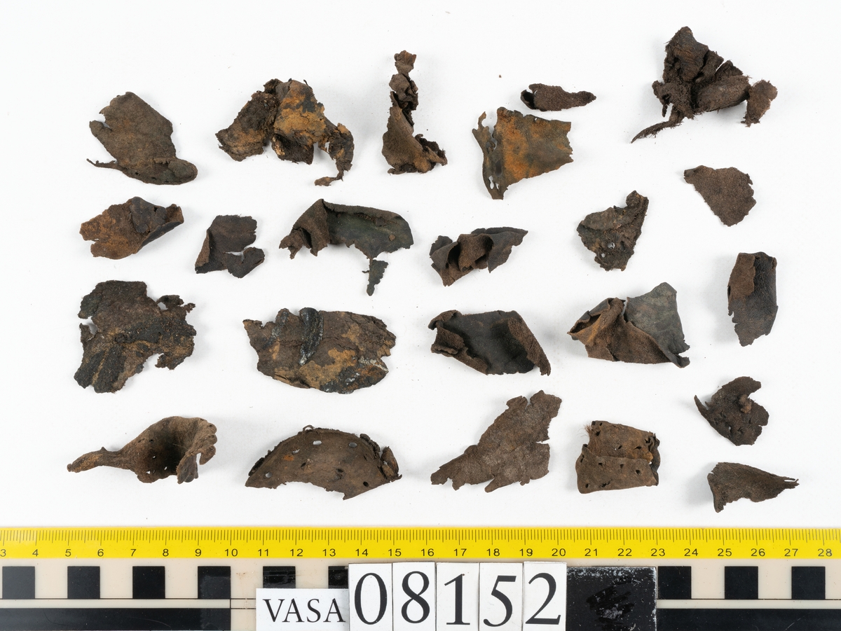 Ett femtiotal större och mindre delar och fragment från två stövlar: sulor, inlägg, ovanläder mm. Lädret ligger vikt. Bland delarna finns också en större klump av tråd eller snöre. Även ulltrådar.

Lädret är hårt och sprött samt har gul-vita utfällningar.