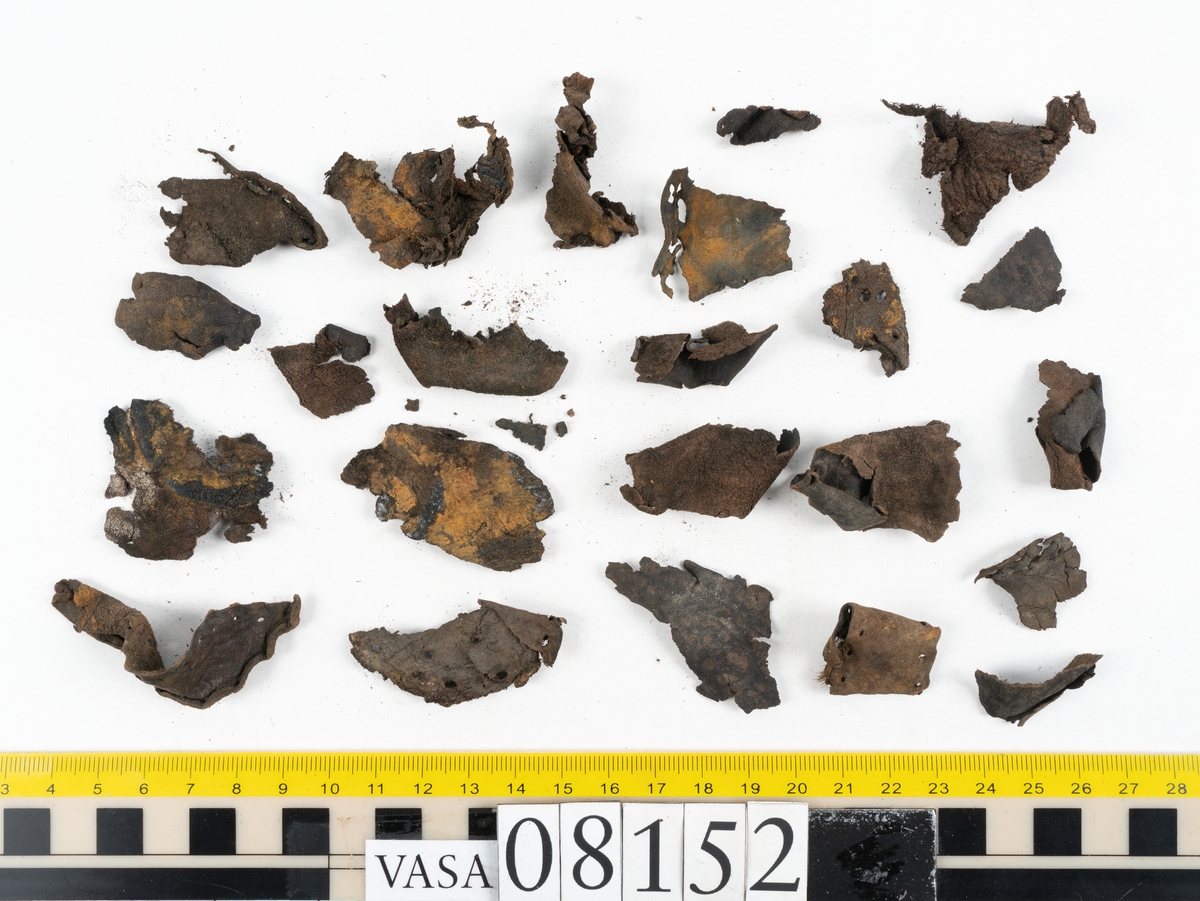 Ett femtiotal större och mindre delar och fragment från två stövlar: sulor, inlägg, ovanläder mm. Lädret ligger vikt. Bland delarna finns också en större klump av tråd eller snöre. Även ulltrådar.

Lädret är hårt och sprött samt har gul-vita utfällningar.