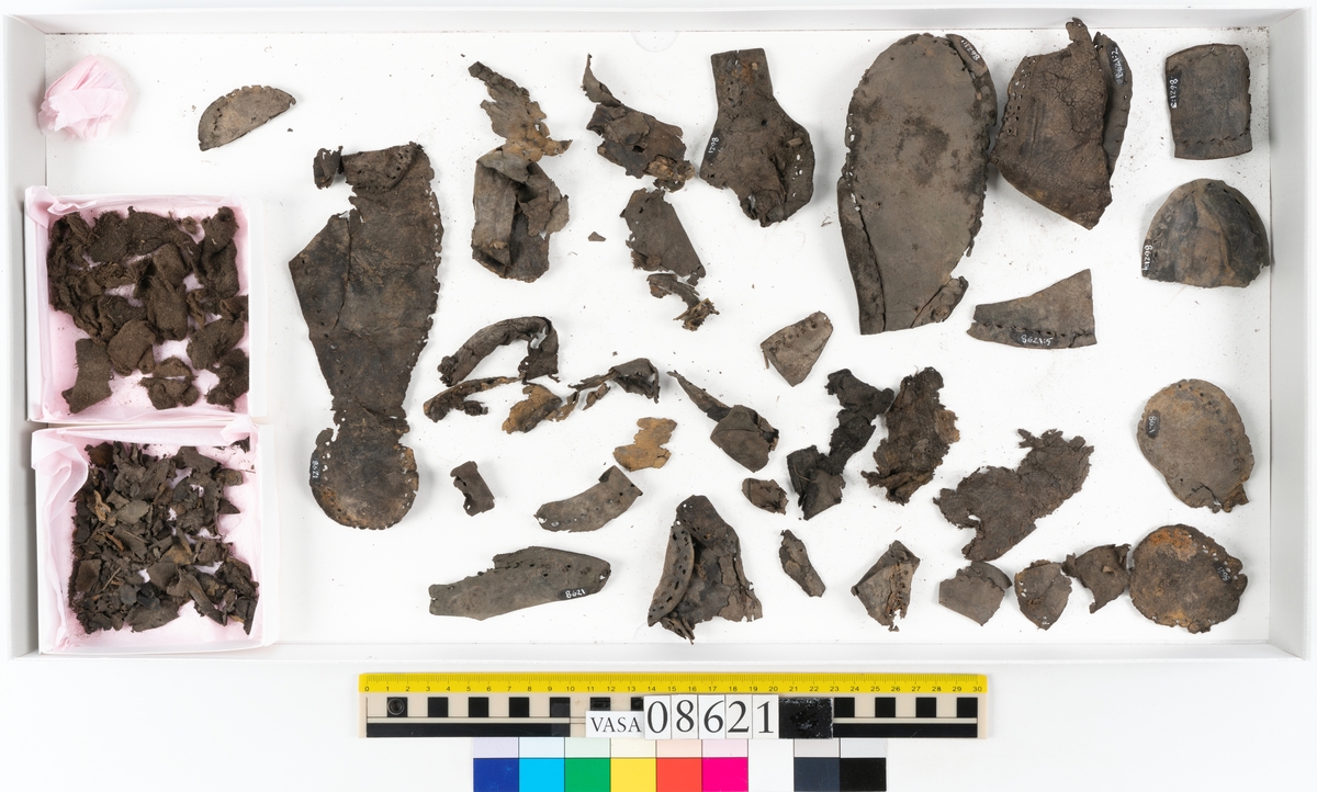 Delar och fragment från sko.
En fragmentarisk sula, fyndnummer 08621. Längs med ytterkanterna löper hål efter stygn. Lädret är spaltat och sprött.
En sula, fyndnummer 08621:1. Sulan är avskuren vid partiet för hålfoten. Sulan har hål efter stygn samt kvarsittande träpligg. Lädret är kraftigt.
En bakkappa, fyndnummer 08621:2. Bakkappan har suttit fästad på skons insida. Längs med ytterkanterna löper stygnshål med kvarsittande tråd.
Två klacklappar, fyndnummer 08621:3 och 08621:4. Klacklapparna har hål efter stygn och/eller träpligg.
Ett inlägg, fyndnummer 08621:5. Lädret är kraftigt.
Ett 100-tal fragment samt ytterligare två klacklappar och två inlägg.
En fragmentarisk bit tråd.
Tidigare även: Ett 15-tal fragment av textil. (Textilen fick ett eget fnr 27904 vid textildokumentationen 2015)
