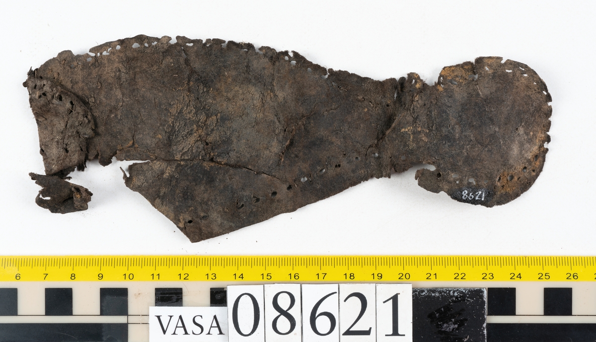 Delar och fragment från sko.
En fragmentarisk sula, fyndnummer 08621. Längs med ytterkanterna löper hål efter stygn. Lädret är spaltat och sprött.
En sula, fyndnummer 08621:1. Sulan är avskuren vid partiet för hålfoten. Sulan har hål efter stygn samt kvarsittande träpligg. Lädret är kraftigt.
En bakkappa, fyndnummer 08621:2. Bakkappan har suttit fästad på skons insida. Längs med ytterkanterna löper stygnshål med kvarsittande tråd.
Två klacklappar, fyndnummer 08621:3 och 08621:4. Klacklapparna har hål efter stygn och/eller träpligg.
Ett inlägg, fyndnummer 08621:5. Lädret är kraftigt.
Ett 100-tal fragment samt ytterligare två klacklappar och två inlägg.
En fragmentarisk bit tråd.
Tidigare även: Ett 15-tal fragment av textil. (Textilen fick ett eget fnr 27904 vid textildokumentationen 2015)