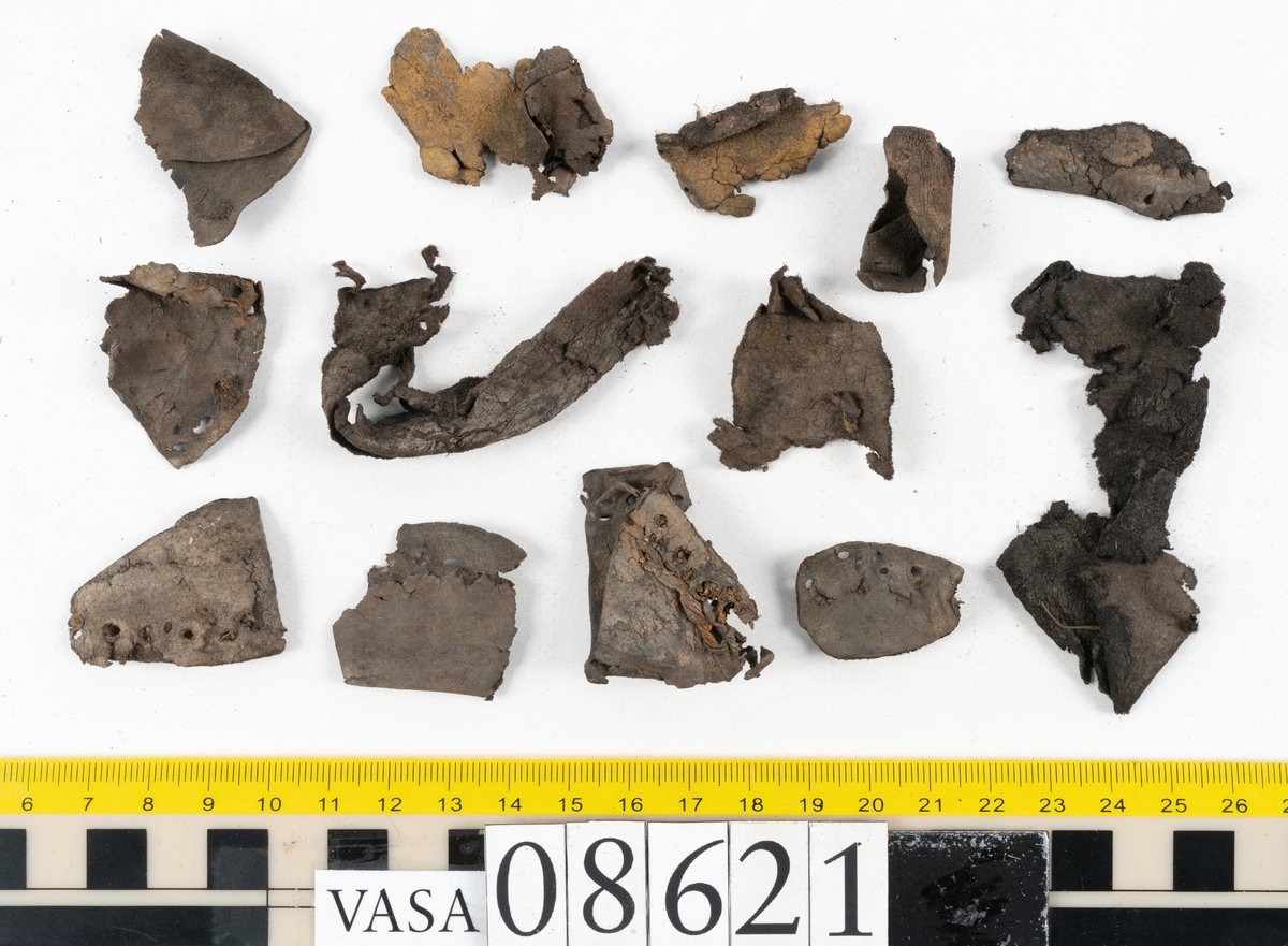 Delar och fragment från sko.
En fragmentarisk sula, fyndnummer 08621. Längs med ytterkanterna löper hål efter stygn. Lädret är spaltat och sprött.
En sula, fyndnummer 08621:1. Sulan är avskuren vid partiet för hålfoten. Sulan har hål efter stygn samt kvarsittande träpligg. Lädret är kraftigt.
En bakkappa, fyndnummer 08621:2. Bakkappan har suttit fästad på skons insida. Längs med ytterkanterna löper stygnshål med kvarsittande tråd.
Två klacklappar, fyndnummer 08621:3 och 08621:4. Klacklapparna har hål efter stygn och/eller träpligg.
Ett inlägg, fyndnummer 08621:5. Lädret är kraftigt.
Ett 100-tal fragment samt ytterligare två klacklappar och två inlägg.
En fragmentarisk bit tråd.
Tidigare även: Ett 15-tal fragment av textil. (Textilen fick ett eget fnr 27904 vid textildokumentationen 2015)