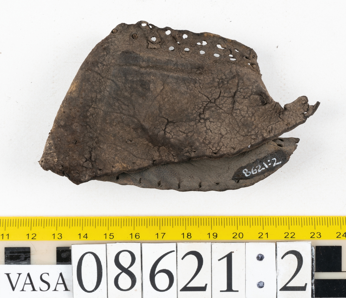 Delar och fragment från sko.
En fragmentarisk sula, fyndnummer 08621. Längs med ytterkanterna löper hål efter stygn. Lädret är spaltat och sprött.
En sula, fyndnummer 08621:1. Sulan är avskuren vid partiet för hålfoten. Sulan har hål efter stygn samt kvarsittande träpligg. Lädret är kraftigt.
En bakkappa, fyndnummer 08621:2. Bakkappan har suttit fästad på skons insida. Längs med ytterkanterna löper stygnshål med kvarsittande tråd.
Två klacklappar, fyndnummer 08621:3 och 08621:4. Klacklapparna har hål efter stygn och/eller träpligg.
Ett inlägg, fyndnummer 08621:5. Lädret är kraftigt.
Ett 100-tal fragment samt ytterligare två klacklappar och två inlägg.
En fragmentarisk bit tråd.
Tidigare även: Ett 15-tal fragment av textil. (Textilen fick ett eget fnr 27904 vid textildokumentationen 2015)