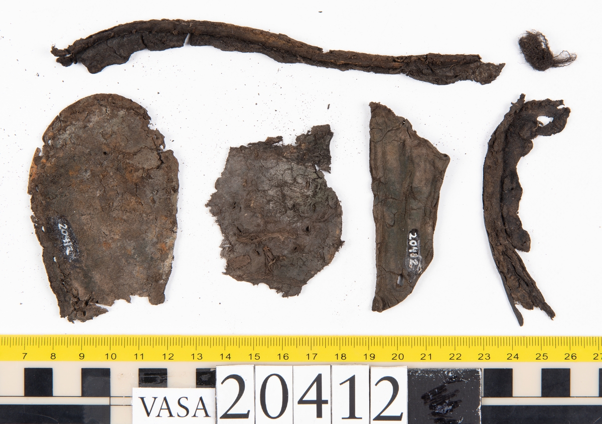 Dokumentation 1997:
Delar och fragment från sko: sulor, slejf, klack, del av holtrand mm. Totalt 23 stycken delar och fragment.
Del av klack bestående av fyra klacklappar. Klacken har kvarsittande skopligg och tråd. Lädret har rostfärgade utfällningar.
Främre partiet av två sulor, sulornas hälparti saknas. Sulorna hålls samman med kvarsittande tråd. Lädret är spaltat.
En fragmentarisk och spaltad sula. Sulan har kvarsittande tråd. Lädret har rostfärgade utfällningar.
En slejf. I slejfens ena ände finns ett snörhål. Runt kanterna löper hål efter stygn.
En textilfiber.

Dokumentation 2022:
Slejf som tillhör bakkappan av sko med fyndnummer 20424.
För mer information, se Uppritning under Länkade filer.

Text in english:
This is the pieced latchet belonging to the shoe quarter 20424.
For more information, go to Länkade filer: Uppritning