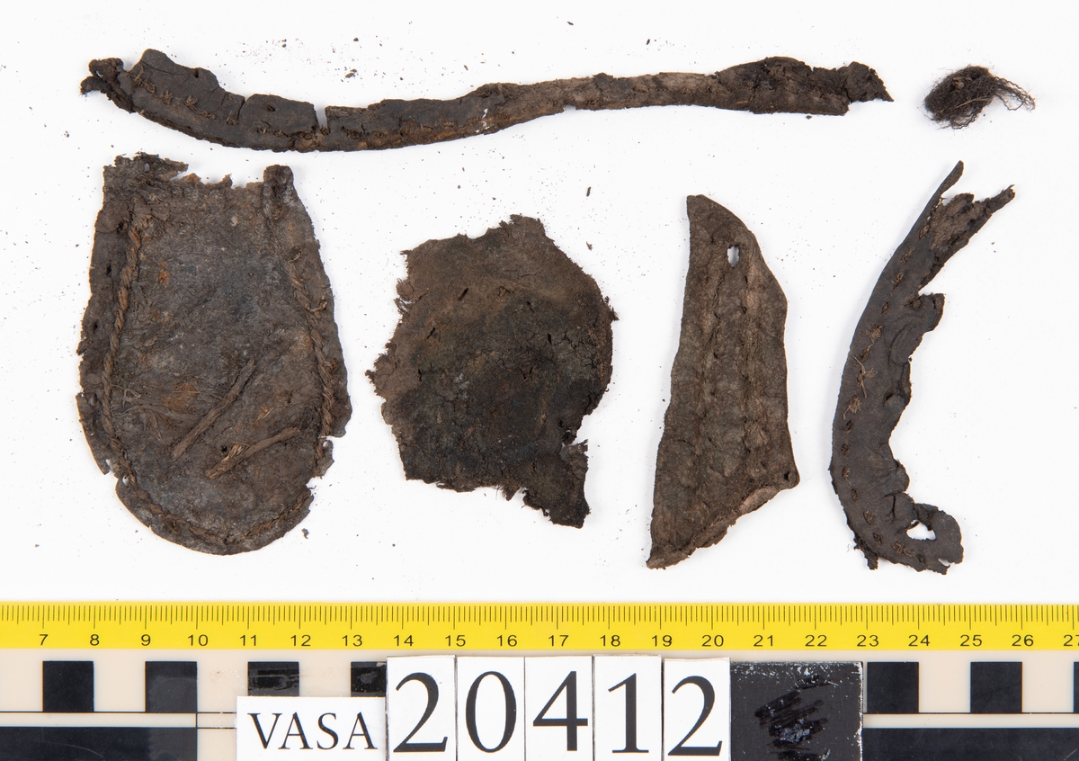 Dokumentation 1997:
Delar och fragment från sko: sulor, slejf, klack, del av holtrand mm. Totalt 23 stycken delar och fragment.
Del av klack bestående av fyra klacklappar. Klacken har kvarsittande skopligg och tråd. Lädret har rostfärgade utfällningar.
Främre partiet av två sulor, sulornas hälparti saknas. Sulorna hålls samman med kvarsittande tråd. Lädret är spaltat.
En fragmentarisk och spaltad sula. Sulan har kvarsittande tråd. Lädret har rostfärgade utfällningar.
En slejf. I slejfens ena ände finns ett snörhål. Runt kanterna löper hål efter stygn.
En textilfiber.

Dokumentation 2022:
Slejf som tillhör bakkappan av sko med fyndnummer 20424.
För mer information, se Uppritning under Länkade filer.

Text in english:
This is the pieced latchet belonging to the shoe quarter 20424.
For more information, go to Länkade filer: Uppritning