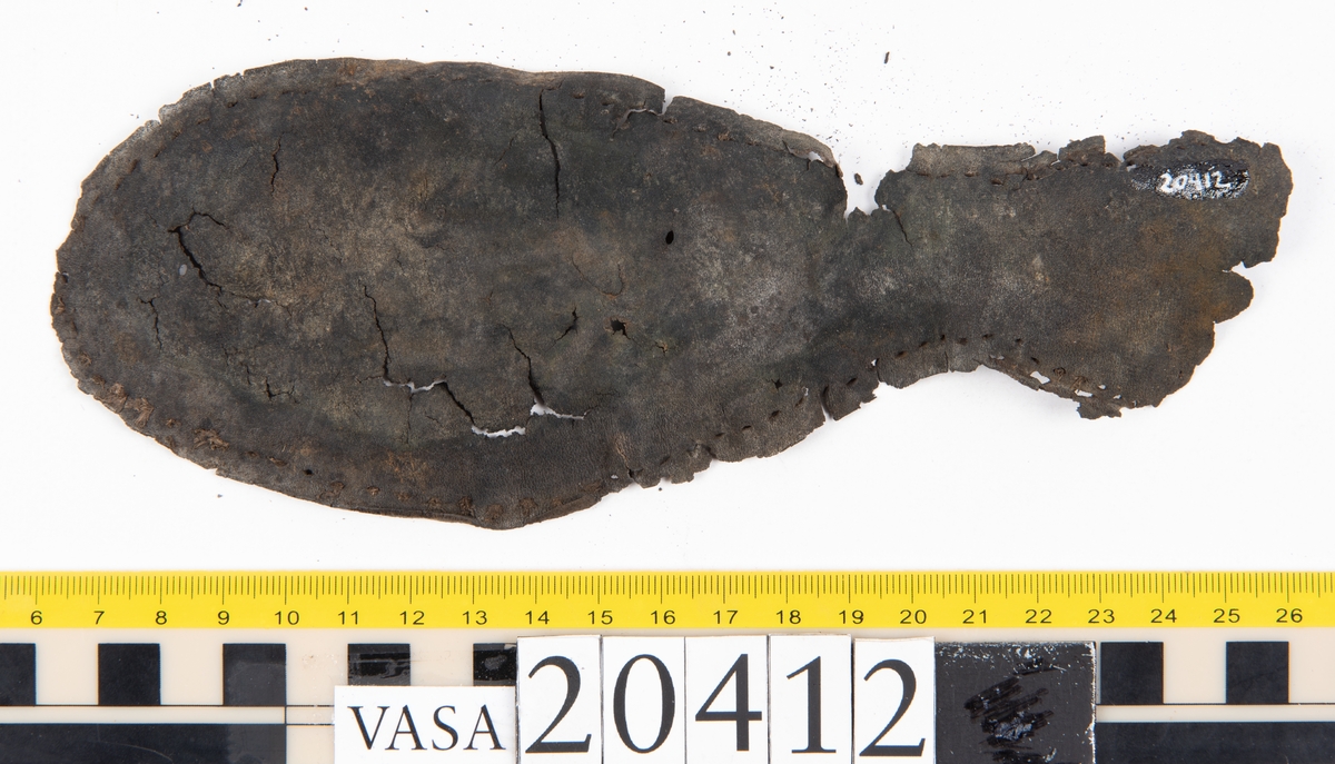 Dokumentation 1997:
Delar och fragment från sko: sulor, slejf, klack, del av holtrand mm. Totalt 23 stycken delar och fragment.
Del av klack bestående av fyra klacklappar. Klacken har kvarsittande skopligg och tråd. Lädret har rostfärgade utfällningar.
Främre partiet av två sulor, sulornas hälparti saknas. Sulorna hålls samman med kvarsittande tråd. Lädret är spaltat.
En fragmentarisk och spaltad sula. Sulan har kvarsittande tråd. Lädret har rostfärgade utfällningar.
En slejf. I slejfens ena ände finns ett snörhål. Runt kanterna löper hål efter stygn.
En textilfiber.

Dokumentation 2022:
Slejf som tillhör bakkappan av sko med fyndnummer 20424.
För mer information, se Uppritning under Länkade filer.

Text in english:
This is the pieced latchet belonging to the shoe quarter 20424.
For more information, go to Länkade filer: Uppritning