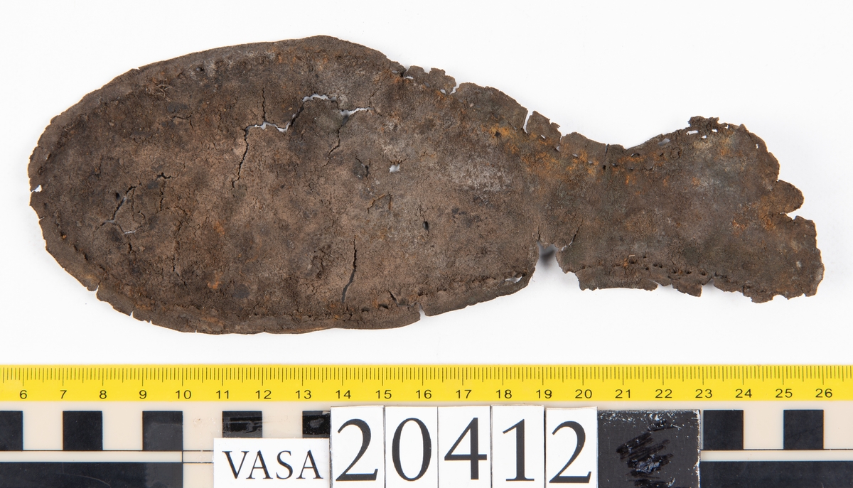 Dokumentation 1997:
Delar och fragment från sko: sulor, slejf, klack, del av holtrand mm. Totalt 23 stycken delar och fragment.
Del av klack bestående av fyra klacklappar. Klacken har kvarsittande skopligg och tråd. Lädret har rostfärgade utfällningar.
Främre partiet av två sulor, sulornas hälparti saknas. Sulorna hålls samman med kvarsittande tråd. Lädret är spaltat.
En fragmentarisk och spaltad sula. Sulan har kvarsittande tråd. Lädret har rostfärgade utfällningar.
En slejf. I slejfens ena ände finns ett snörhål. Runt kanterna löper hål efter stygn.
En textilfiber.

Dokumentation 2022:
Slejf som tillhör bakkappan av sko med fyndnummer 20424.
För mer information, se Uppritning under Länkade filer.

Text in english:
This is the pieced latchet belonging to the shoe quarter 20424.
For more information, go to Länkade filer: Uppritning