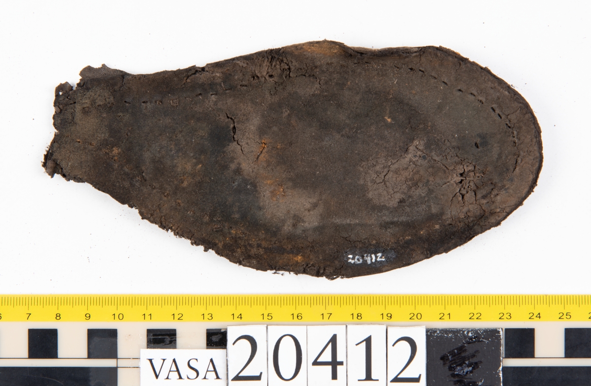 Dokumentation 1997:
Delar och fragment från sko: sulor, slejf, klack, del av holtrand mm. Totalt 23 stycken delar och fragment.
Del av klack bestående av fyra klacklappar. Klacken har kvarsittande skopligg och tråd. Lädret har rostfärgade utfällningar.
Främre partiet av två sulor, sulornas hälparti saknas. Sulorna hålls samman med kvarsittande tråd. Lädret är spaltat.
En fragmentarisk och spaltad sula. Sulan har kvarsittande tråd. Lädret har rostfärgade utfällningar.
En slejf. I slejfens ena ände finns ett snörhål. Runt kanterna löper hål efter stygn.
En textilfiber.

Dokumentation 2022:
Slejf som tillhör bakkappan av sko med fyndnummer 20424.
För mer information, se Uppritning under Länkade filer.

Text in english:
This is the pieced latchet belonging to the shoe quarter 20424.
For more information, go to Länkade filer: Uppritning