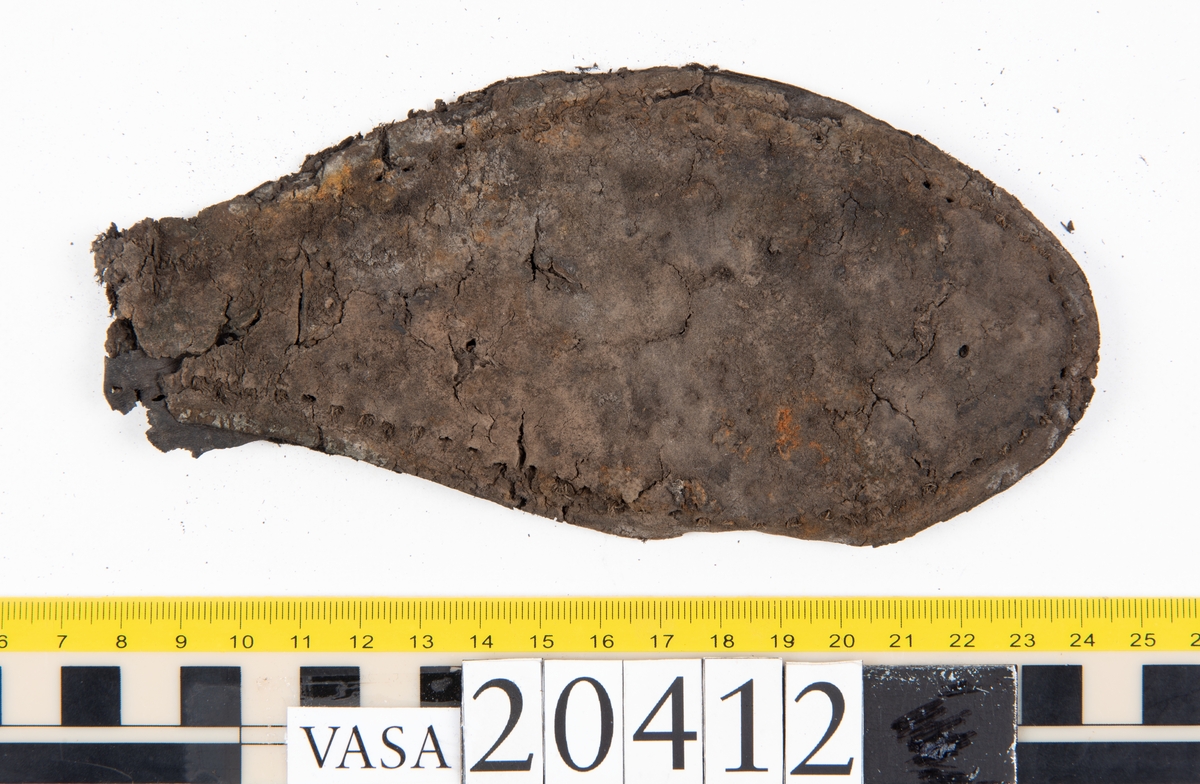 Dokumentation 1997:
Delar och fragment från sko: sulor, slejf, klack, del av holtrand mm. Totalt 23 stycken delar och fragment.
Del av klack bestående av fyra klacklappar. Klacken har kvarsittande skopligg och tråd. Lädret har rostfärgade utfällningar.
Främre partiet av två sulor, sulornas hälparti saknas. Sulorna hålls samman med kvarsittande tråd. Lädret är spaltat.
En fragmentarisk och spaltad sula. Sulan har kvarsittande tråd. Lädret har rostfärgade utfällningar.
En slejf. I slejfens ena ände finns ett snörhål. Runt kanterna löper hål efter stygn.
En textilfiber.

Dokumentation 2022:
Slejf som tillhör bakkappan av sko med fyndnummer 20424.
För mer information, se Uppritning under Länkade filer.

Text in english:
This is the pieced latchet belonging to the shoe quarter 20424.
For more information, go to Länkade filer: Uppritning